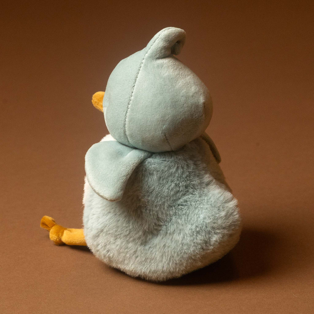 A plush roly-poly-birdie-tweet with pale blue feathers, a white belly, and orange beak and legs shown from the back on a solid brown background. The toy is light blue-gray with soft yellow accents on its beak and feet.