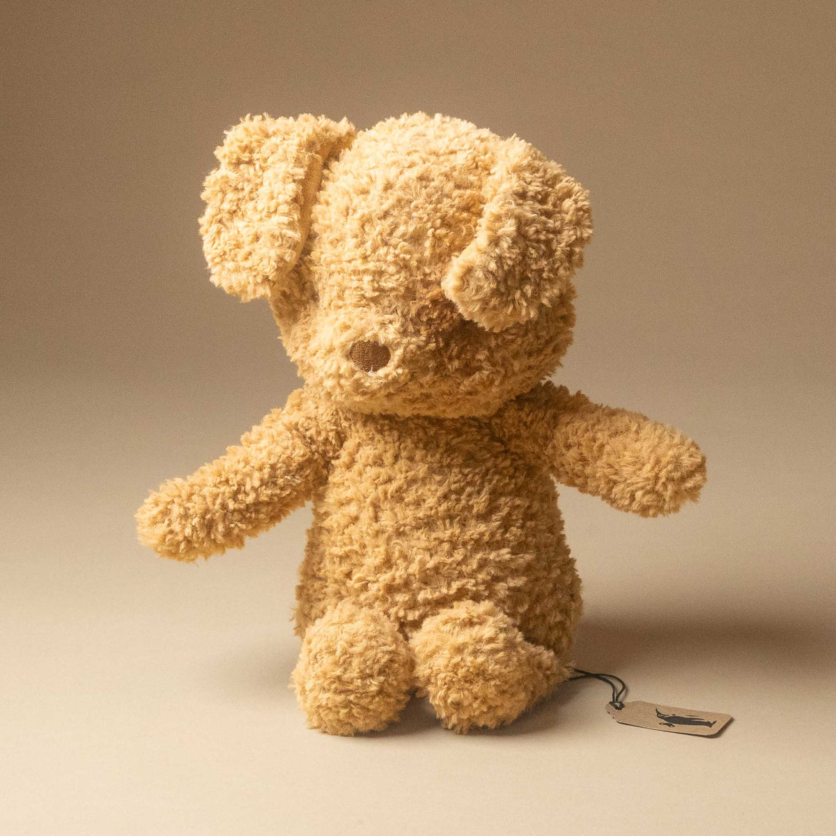 rufus-the-dog in a curly, bouclé-like tan fabric, photographed against a warm beige studio background. A small hangtag is attached near the right side of the bear.