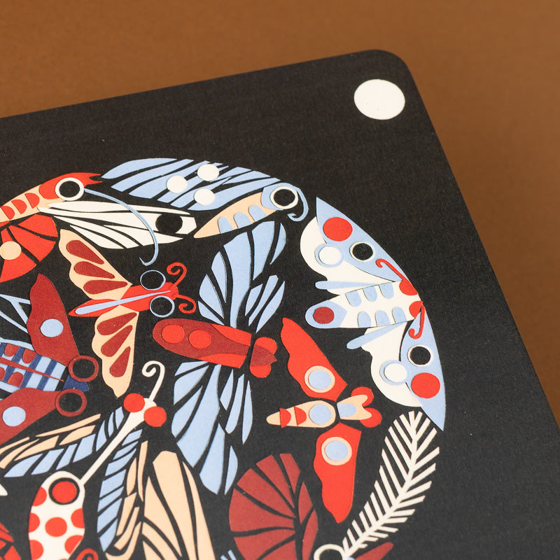 A close-up of a black rectangular product, likely a tablet case or notebook cover, featuring a colorful retro insect and leaf pattern in red, light blue, beige, and white. The product is shown resting on a solid brown background.
