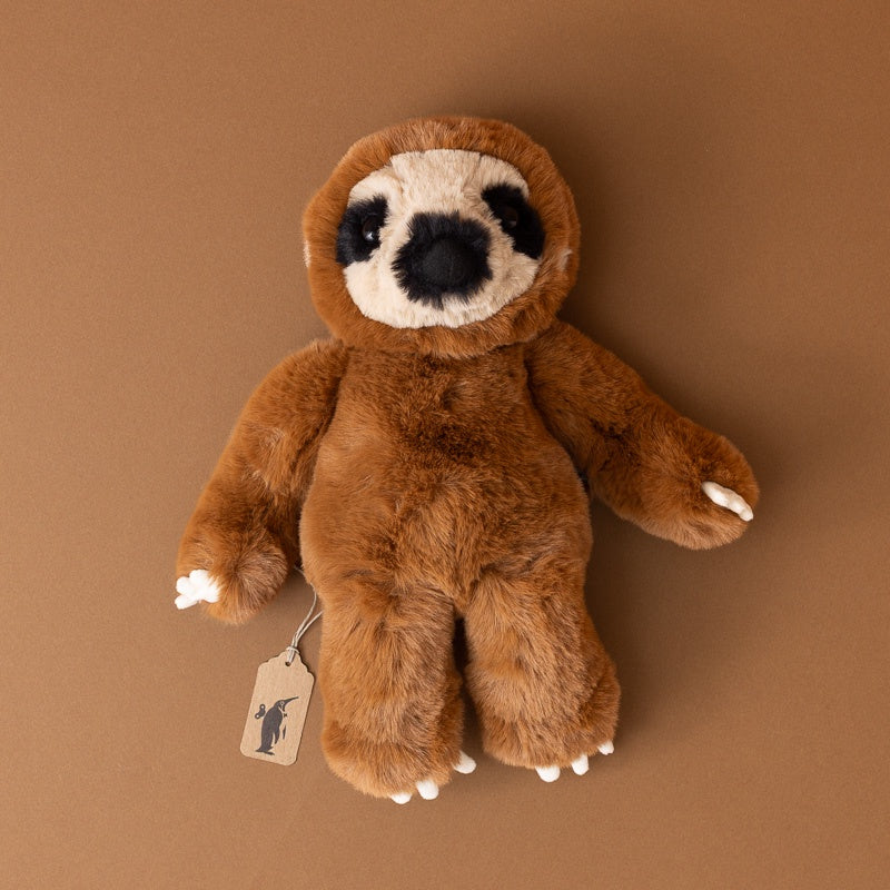 A plush sloth toy with brown fur, a cream-colored snout and shoulders, and black facial features, photographed on a solid brown background. A small paper tag hangs from one hand.