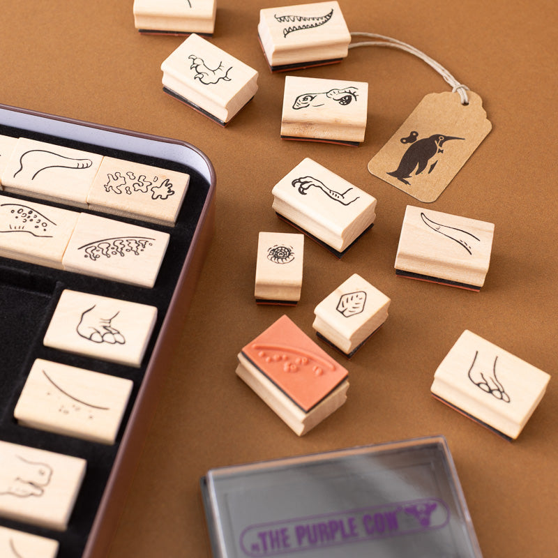 A set of rubber stamp blocks with engraved animal and nature designs, displayed on a brown surface alongside a metal tray, a kraft paper tag, and a purple ink pad