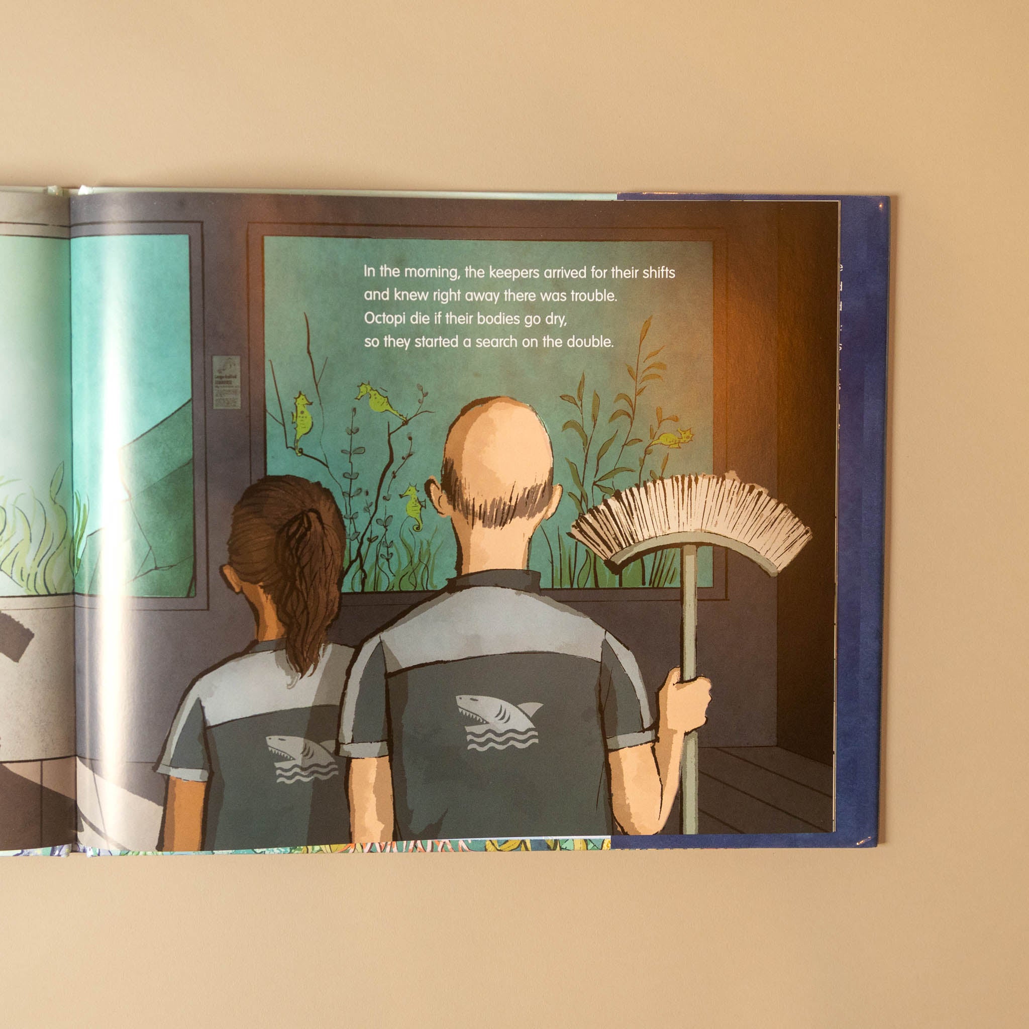 An open children's book or illustrated print showing an illustration of two people from behind in a poolside setting, with text on the page, photographed against a beige wall.