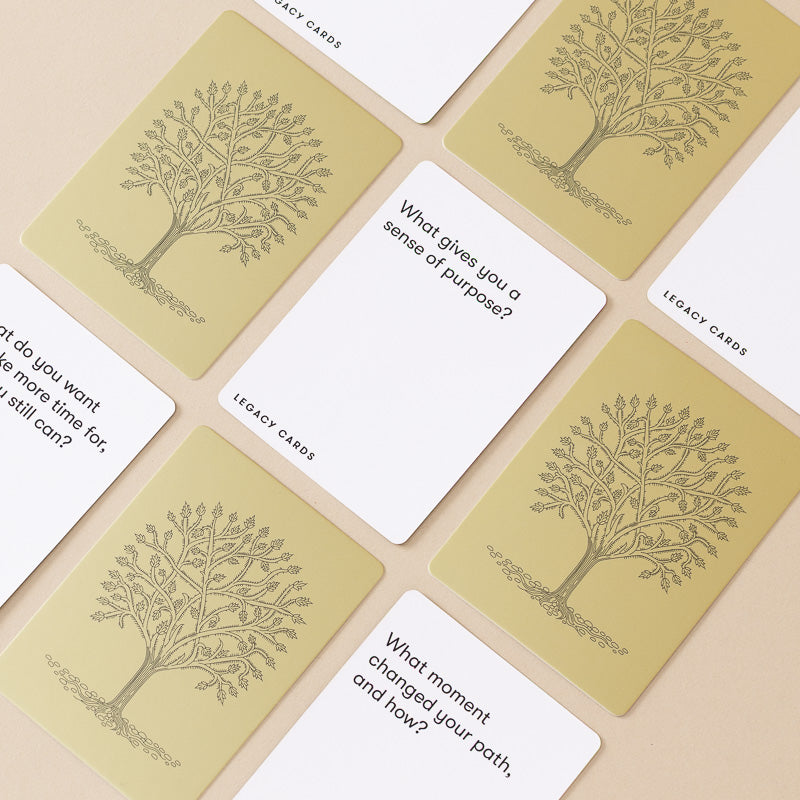 A set of reflective legacy cards arranged in a flat-lay pattern on a light beige background. The cards feature a warm beige back with a printed tree illustration and the text 'LEGACY CARDS,' while the front faces are white with black text prompts such as 'What gives you a sense of purpose?' and 'What moment changed your path, and how?'.