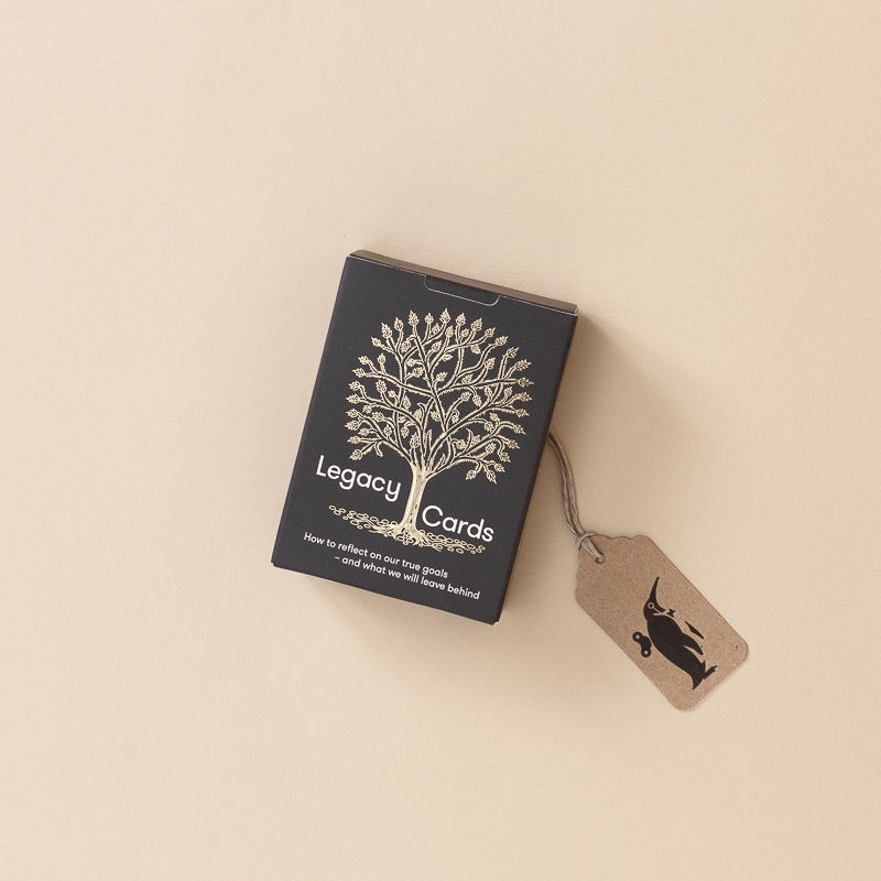 A black box of Legacy Cards with a white tree illustration and text, placed on a warm beige background with a small kraft paper tag attached by a string.