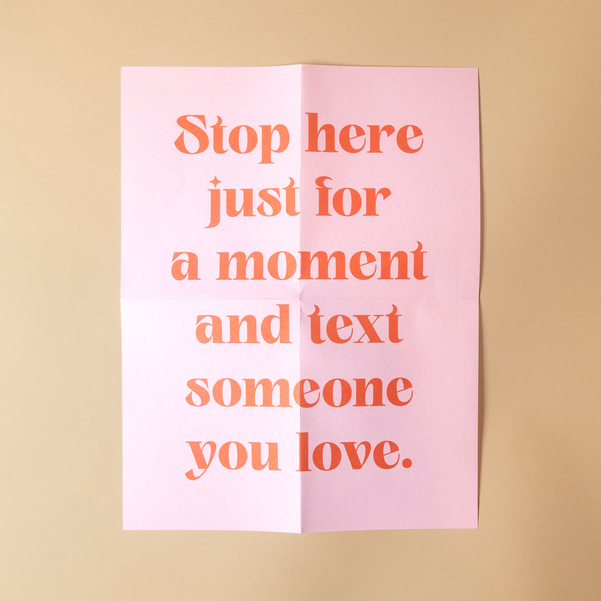 A pink folded paper card with orange text reading 'Stop here just for a moment and text someone you love.' on a warm beige background.