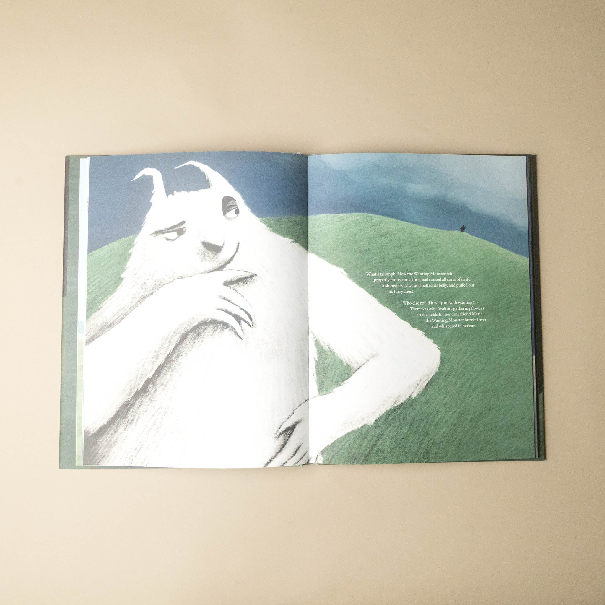 An open children's book laid flat on a beige surface, showing a surreal illustration of a white goat-like creature on a green hill under a blue sky, with small printed text on the right page.