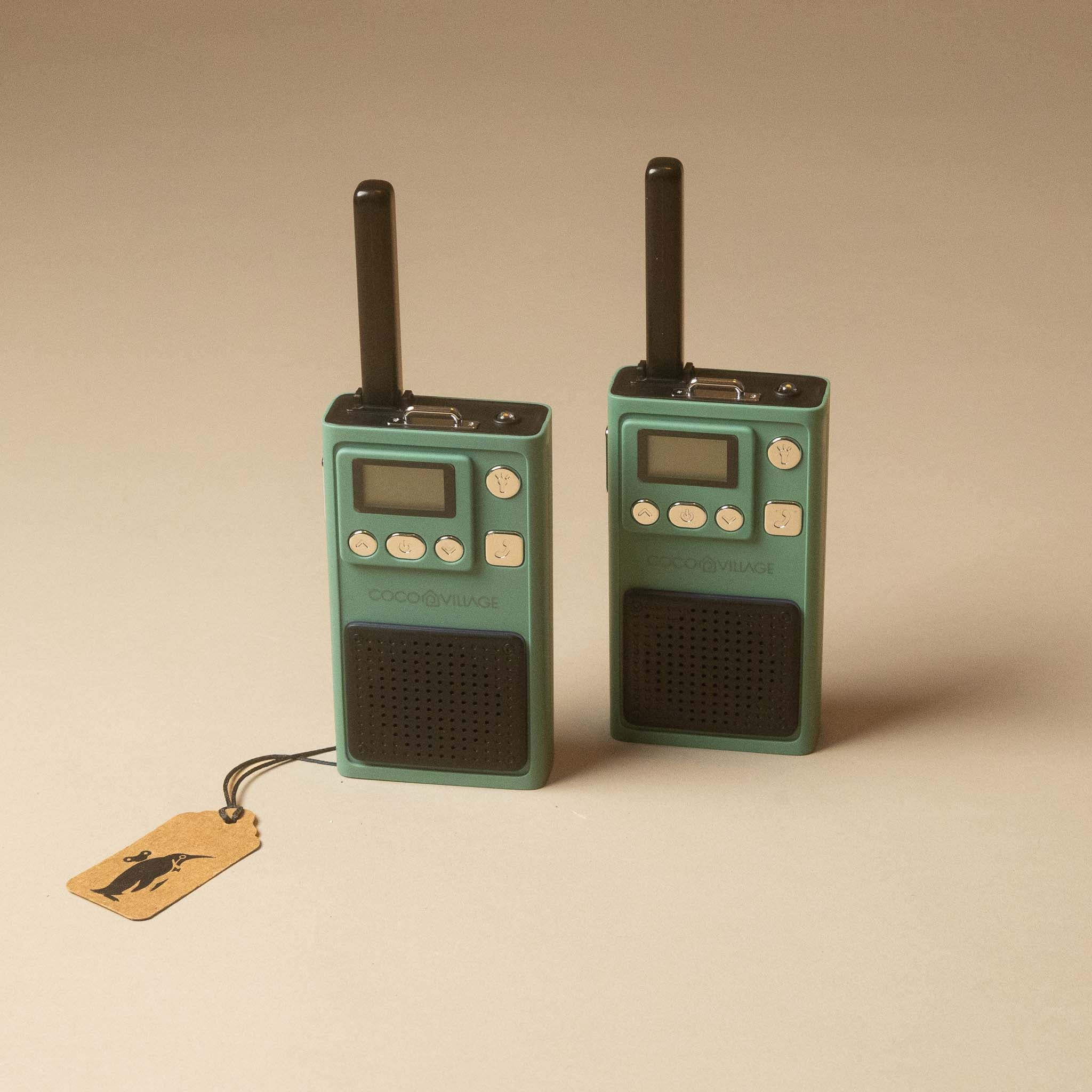 Two retro-style walkie-talkies in mint green and black, standing upright on a warm beige background. One has a small attached kraft paper tag with a black logo.