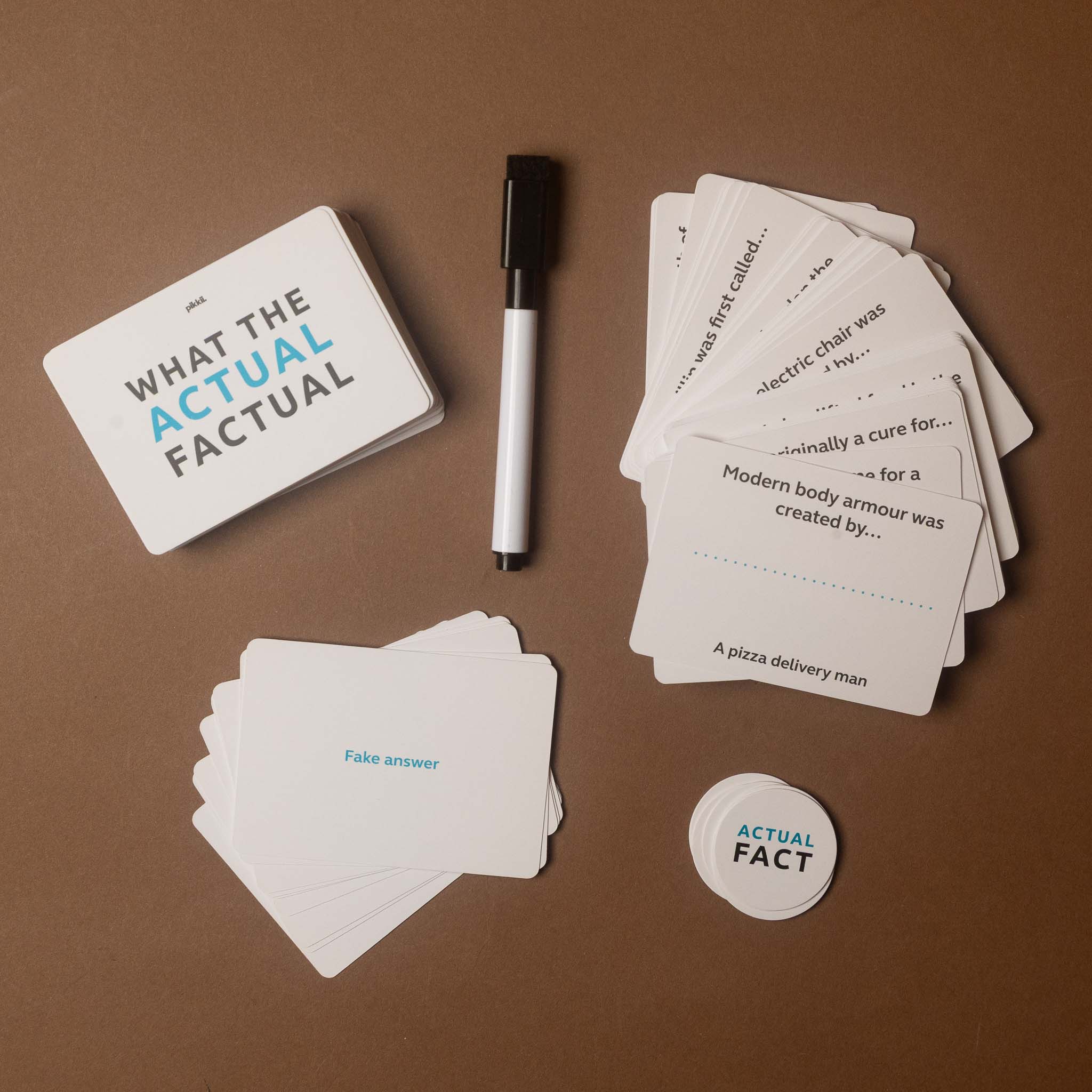 A boxed card game set labeled 'WHAT THE ACTUAL FACTUAL' arranged on a brown surface with a black marker, a stack of white cards, a small round playing piece, and a few sample cards showing humorous fake-answer questions.