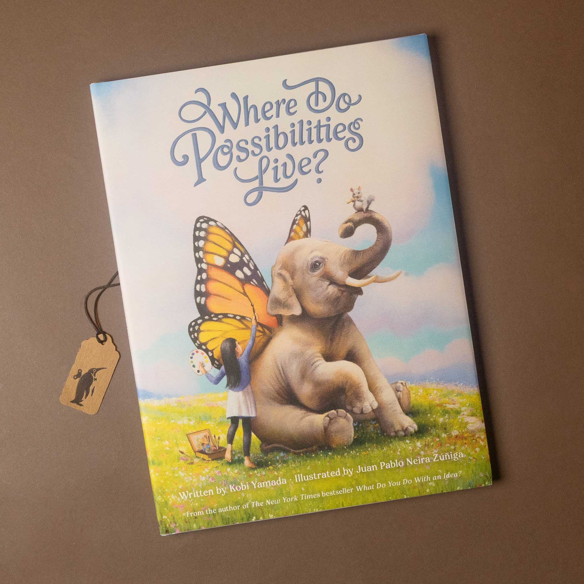 Children's book titled "Where Do Possibilities Live?" shown at an angle on a brown background, with a small hanging tag beside it. The cover features an illustrated elephant with butterfly wings and a child standing beside it.