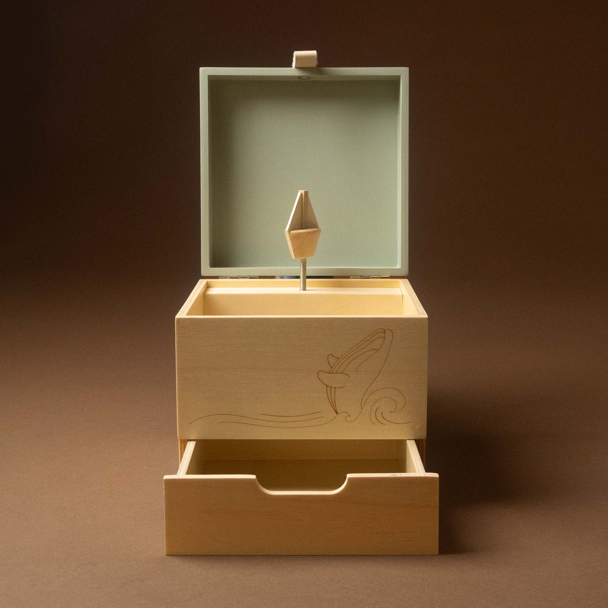 wooden-musical-treasures-box-by-the-sea with an open top lid and a pulled-out drawer, shown on a dark brown studio background. The front of the box has a light engraved wave and fish design.