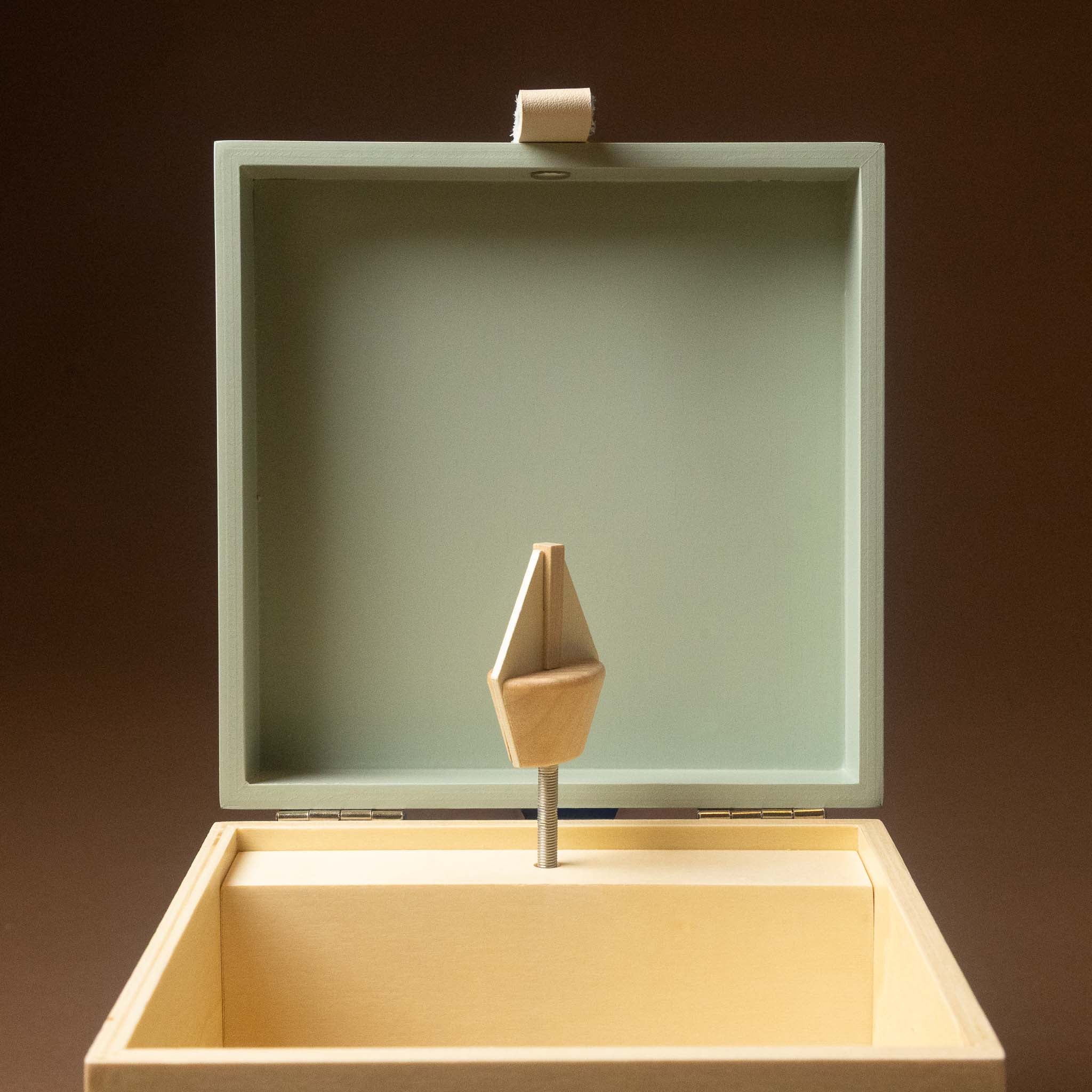 wooden-musical-treasures-box-by-the-sea with an open lid, featuring a pale sage green interior and a light natural wood turnkey or spool-like handle mounted on a metal rod inside. The box is photographed against a dark brown studio background.