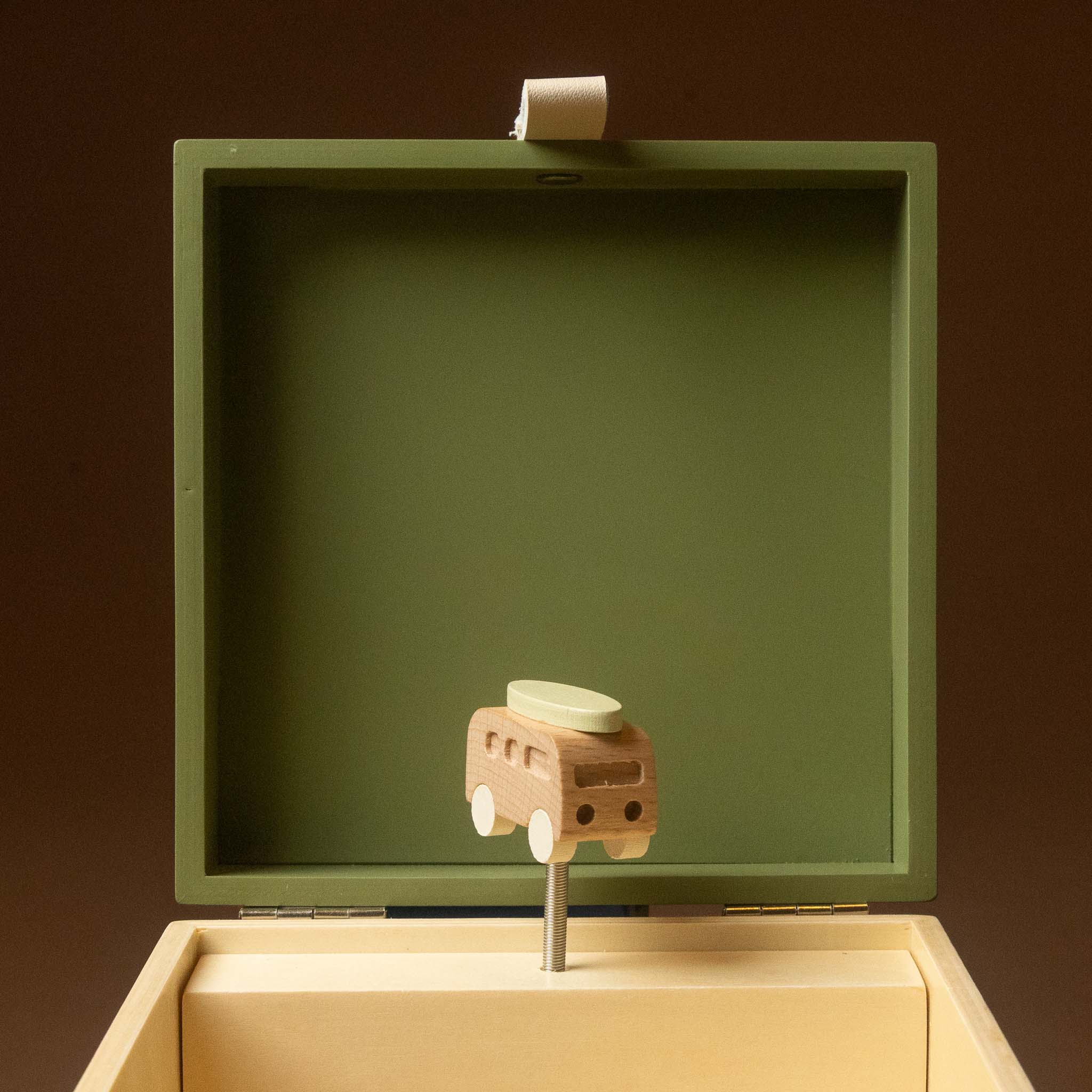 wooden-musical-treasures-box-road-warrier with wooden toy bus with a light green roof sits on a metal stand inside an open cream-colored music box with an olive green lid, set against a dark brown background.