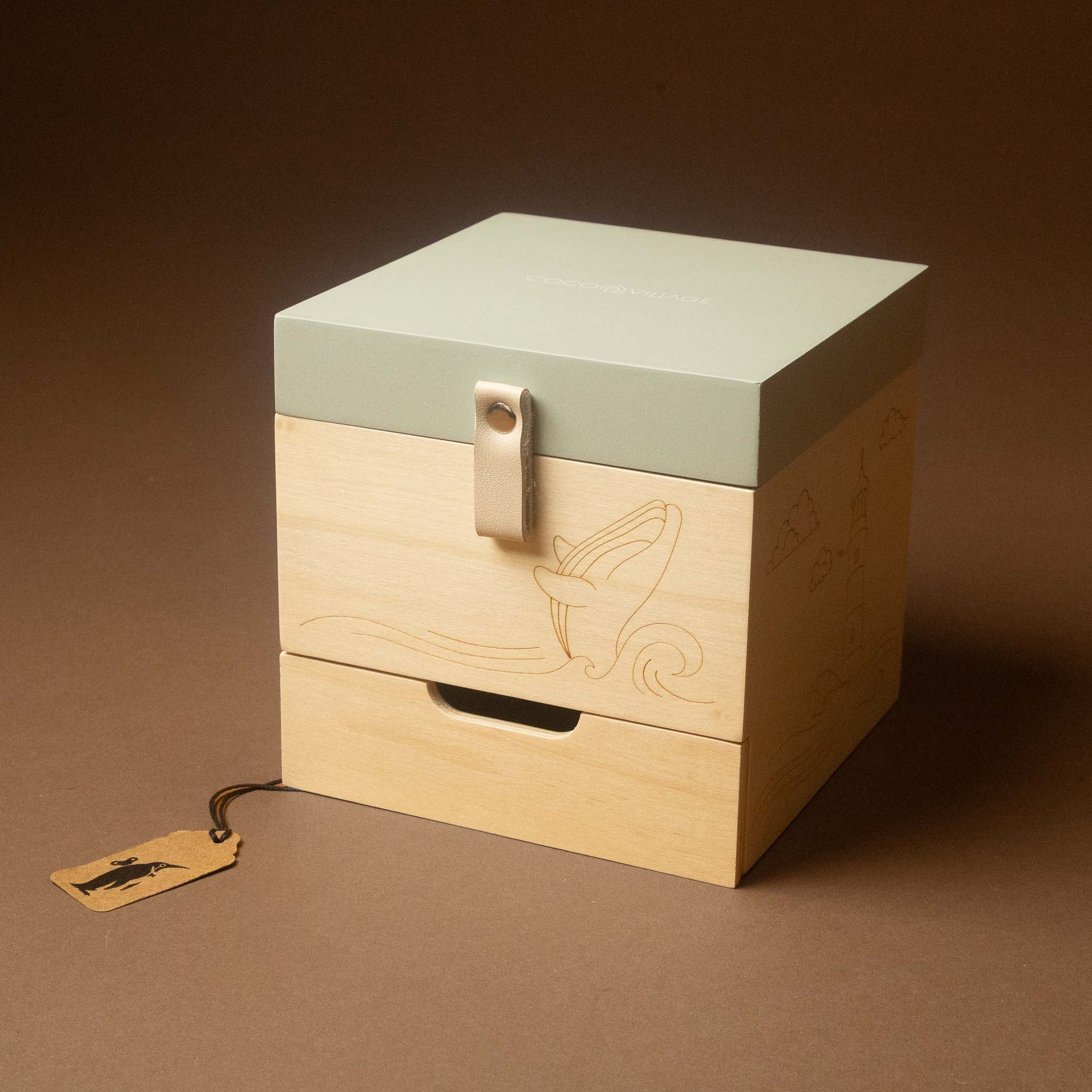 wooden-musical-treasures-box-by-the-sea with a pale sage green top lid and two light natural wood drawers, photographed on a warm brown studio background. A small hanging tag is attached to the front drawer.