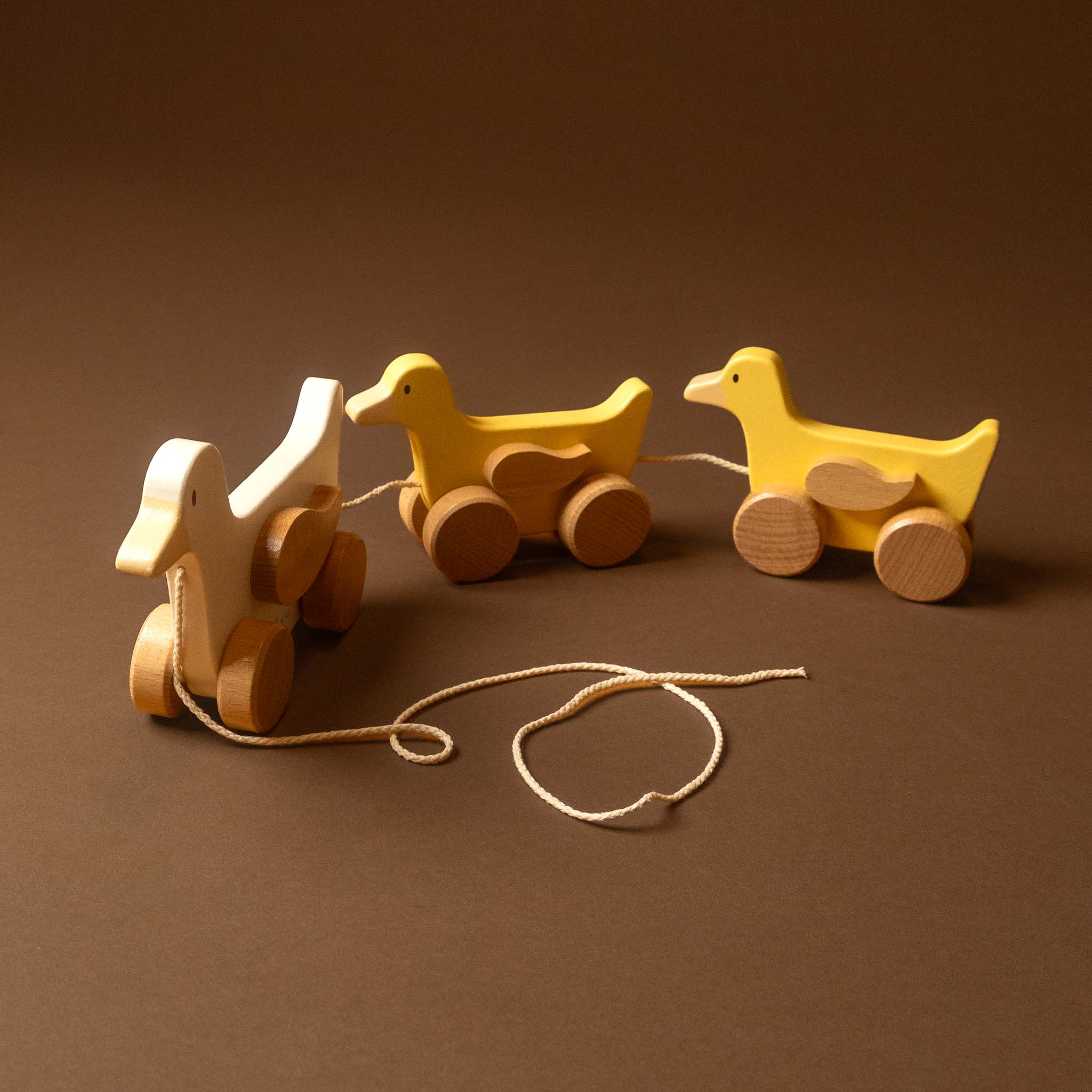 Three wooden toy ducks with wheels on a brown background. One duck is natural wood, and two are painted yellow, connected by a rope.