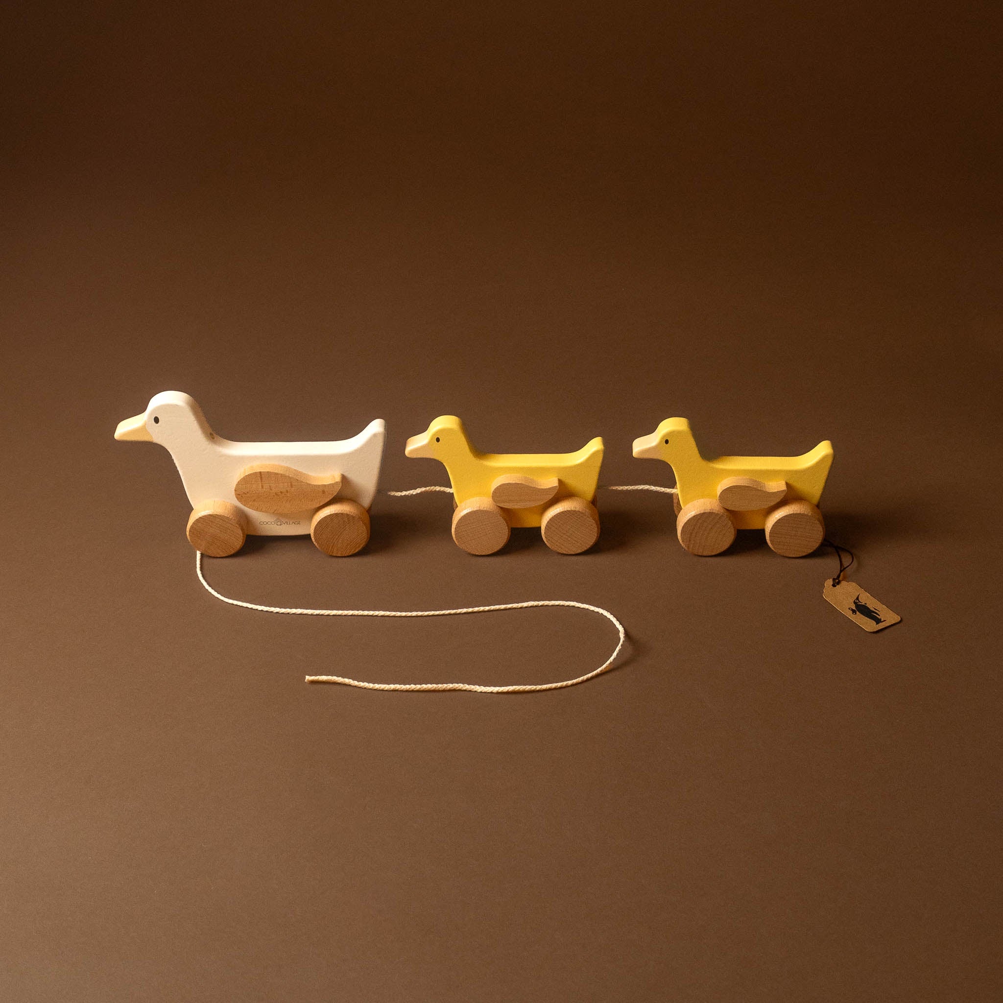 Three wooden toy ducks with wheels, including one larger white duck pulling two smaller yellow ducks by a string, photographed on a solid brown background.