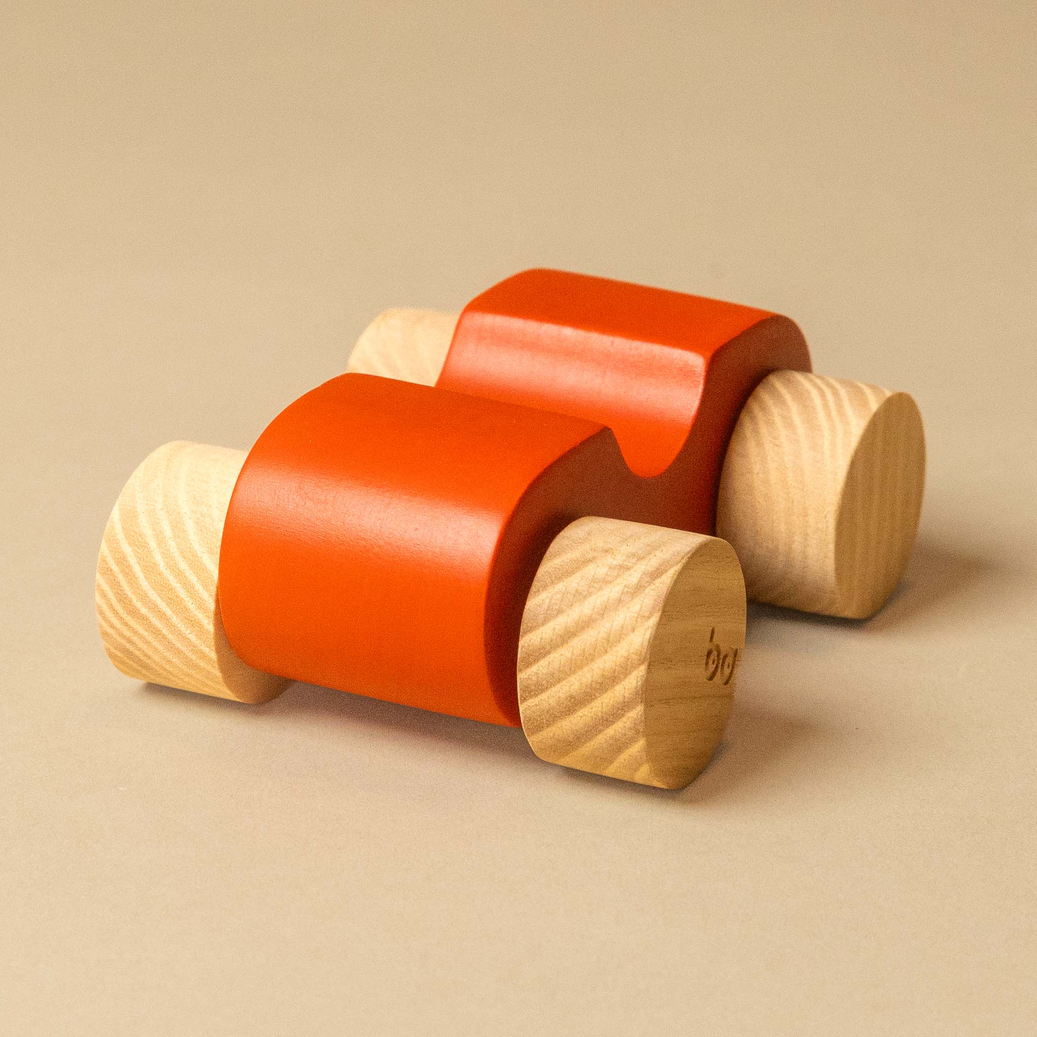A wooden toy car with a bright orange-red painted body and natural light wood wheels, photographed on a plain beige background.