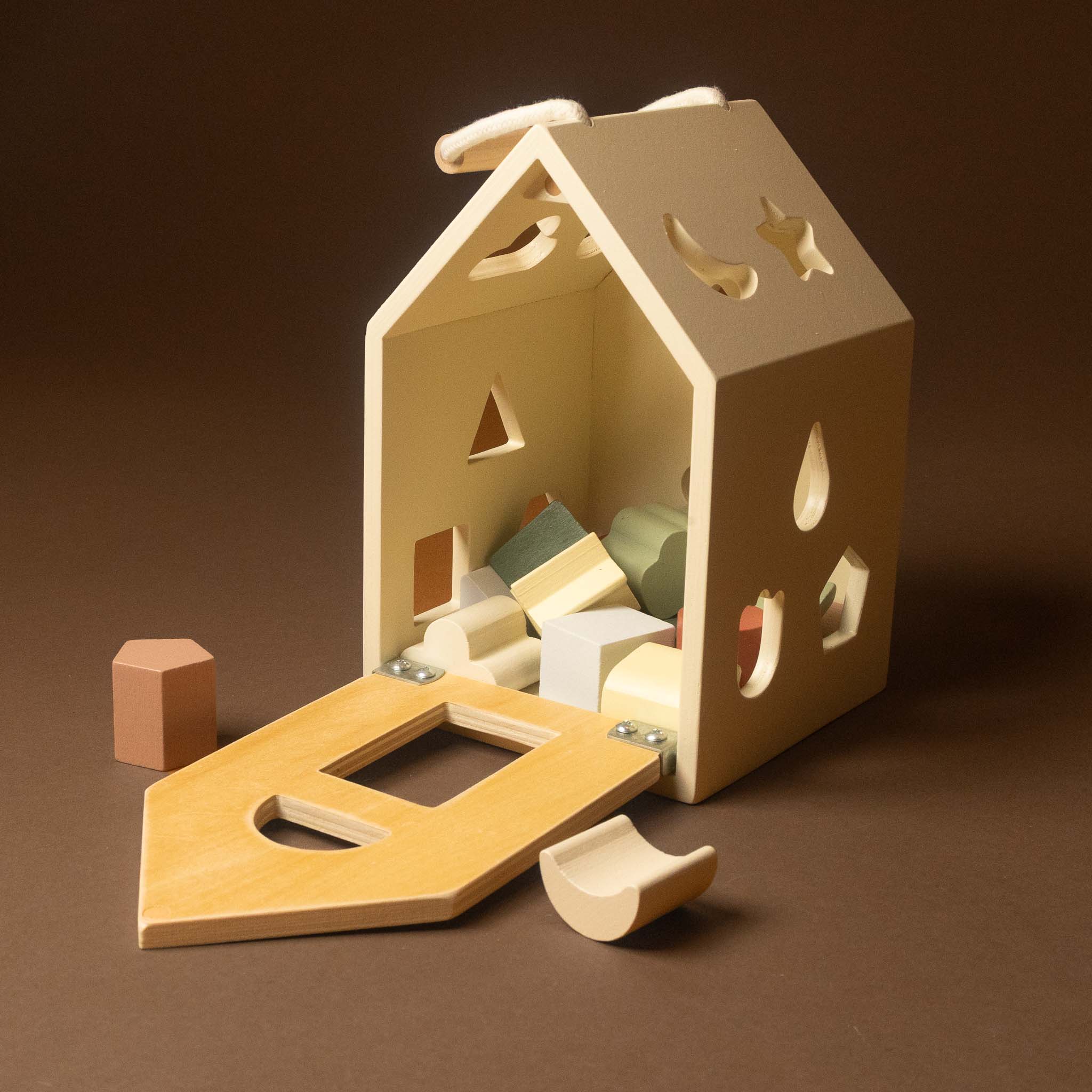 A wooden house-shaped toy with a sliding open door and cutout details, filled with small geometric blocks, photographed on a dark brown studio background.