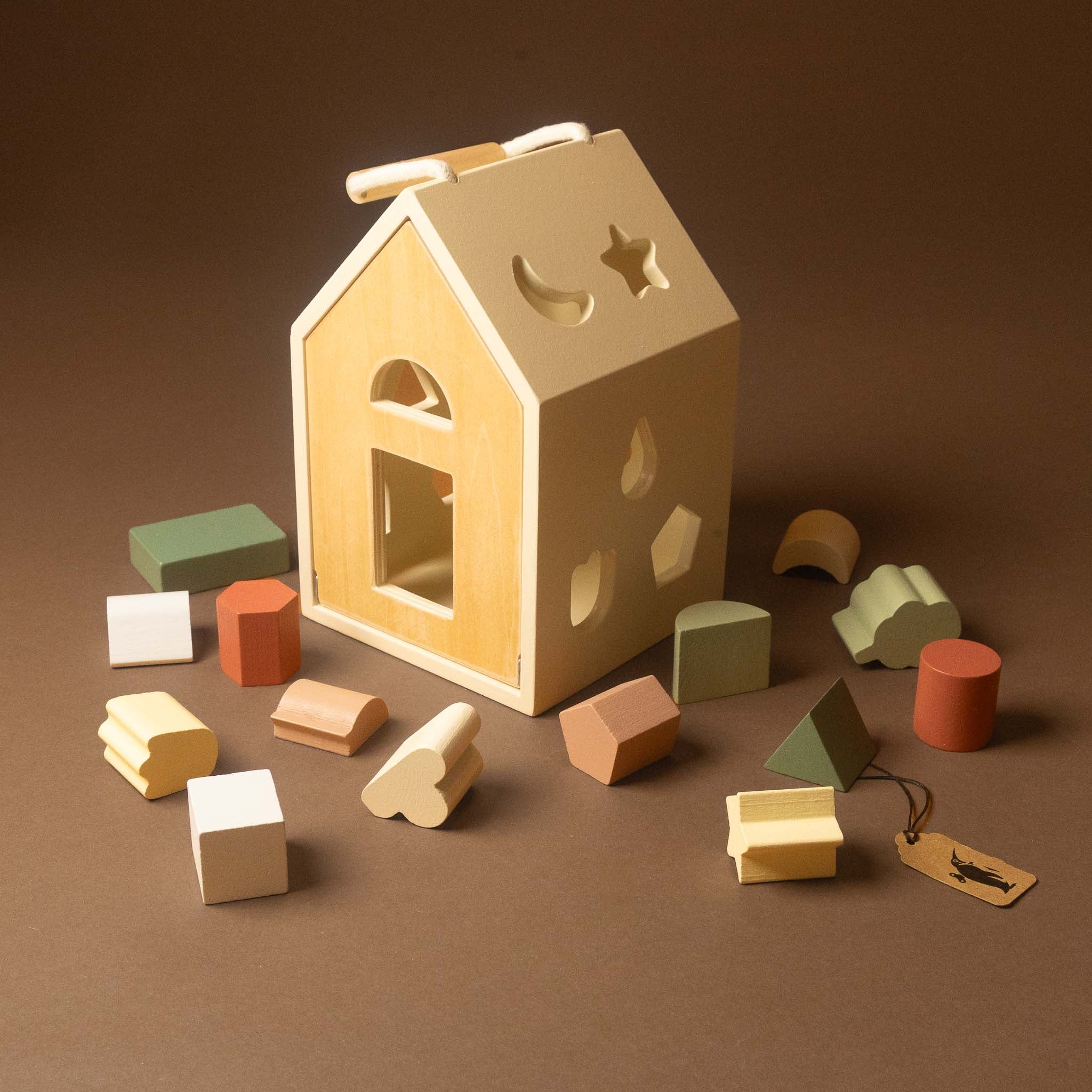 A wooden house-shaped shape-sorting toy with cutout windows and geometric blocks in soft pastel colors arranged around it on a warm brown studio background.