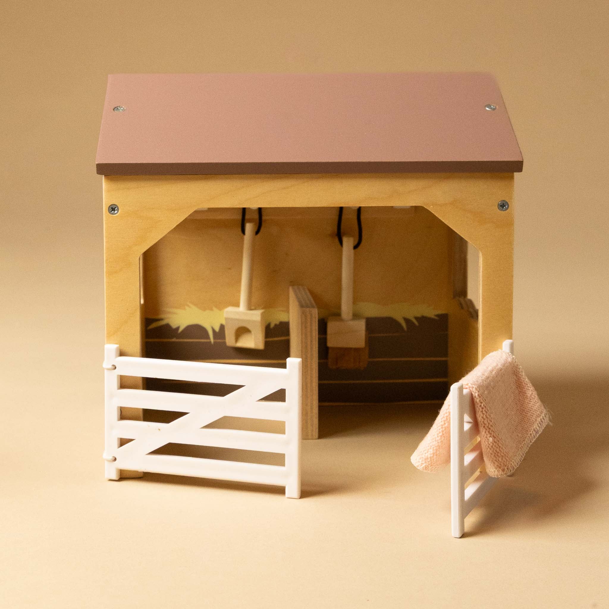 Wooden wooden-stable-play-set with a pink roof, white gate, and pink towel on a beige background