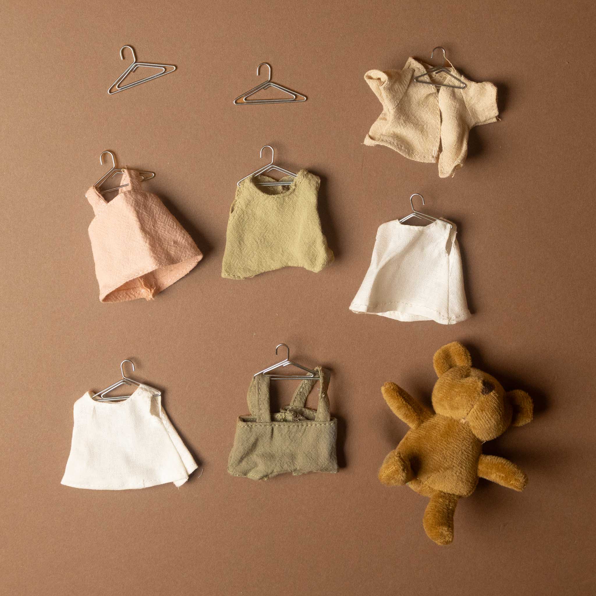 woodland-bears-wardrobe miniature clothing and accessories displayed on a warm brown background, including several small white and beige garments on hangers, a peach sleeveless top, an olive green sleeveless top, a cream sleeveless top, a green pair of mini overalls, and a brown plush teddy bear.