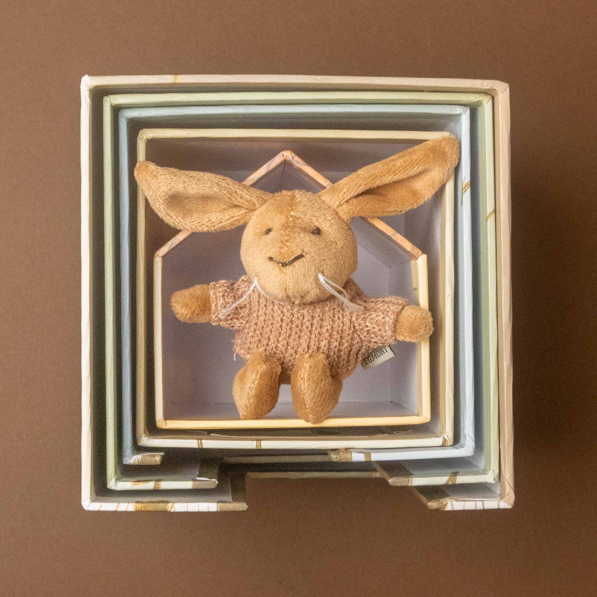 A small plush bunny toy with long tan ears and a knitted pink sweater, displayed inside a woodland-friends-nesting-blocks-house-lenny-rabbit with layered pastel blue, green, and cream borders against a brown background.