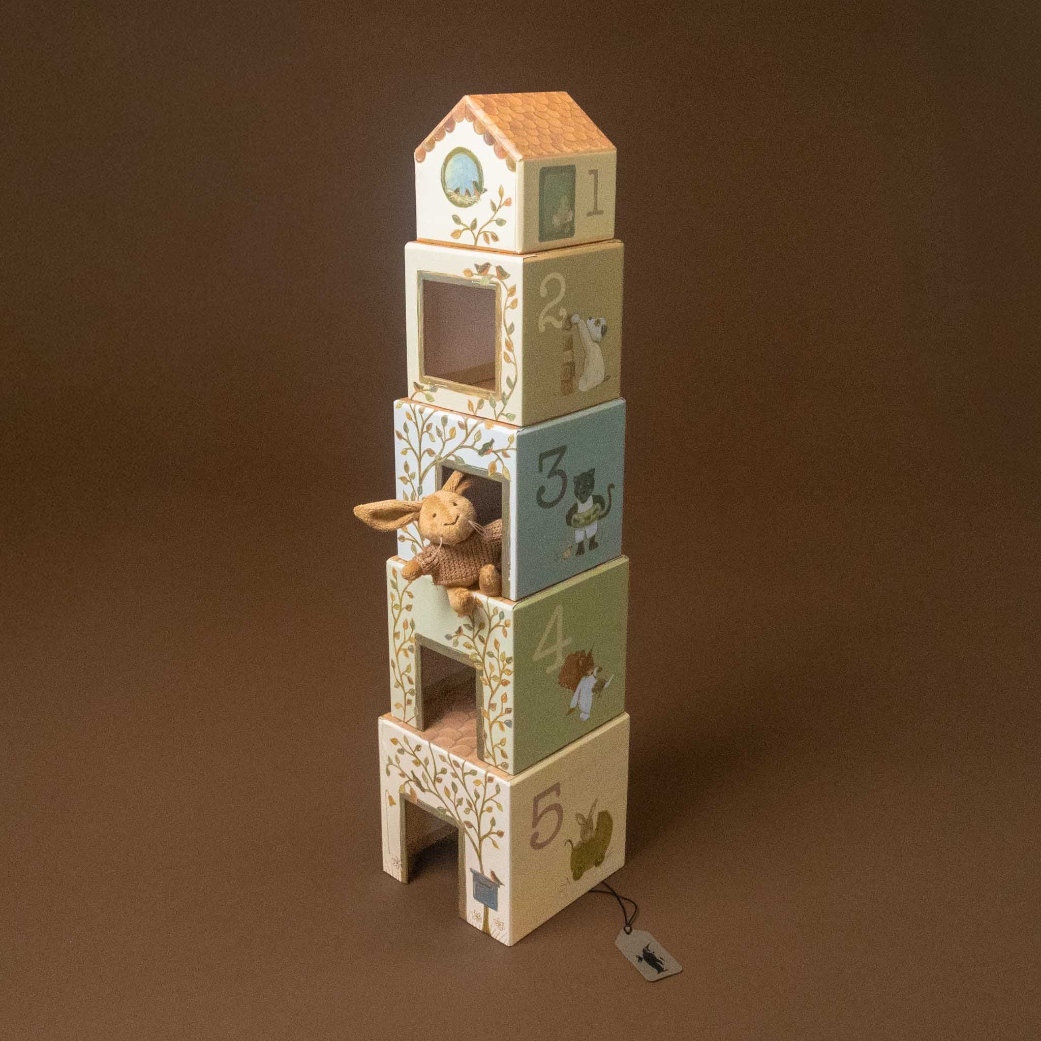 woodland-friends-nesting-blocks-house-lenny-rabbit A wooden toy house block tower with five stacked numbered boxes in soft pastel colors, decorated with floral patterns and animal illustrations, with a small stuffed bunny tucked into the middle box. A small product tag hangs from the base against a solid brown studio background.