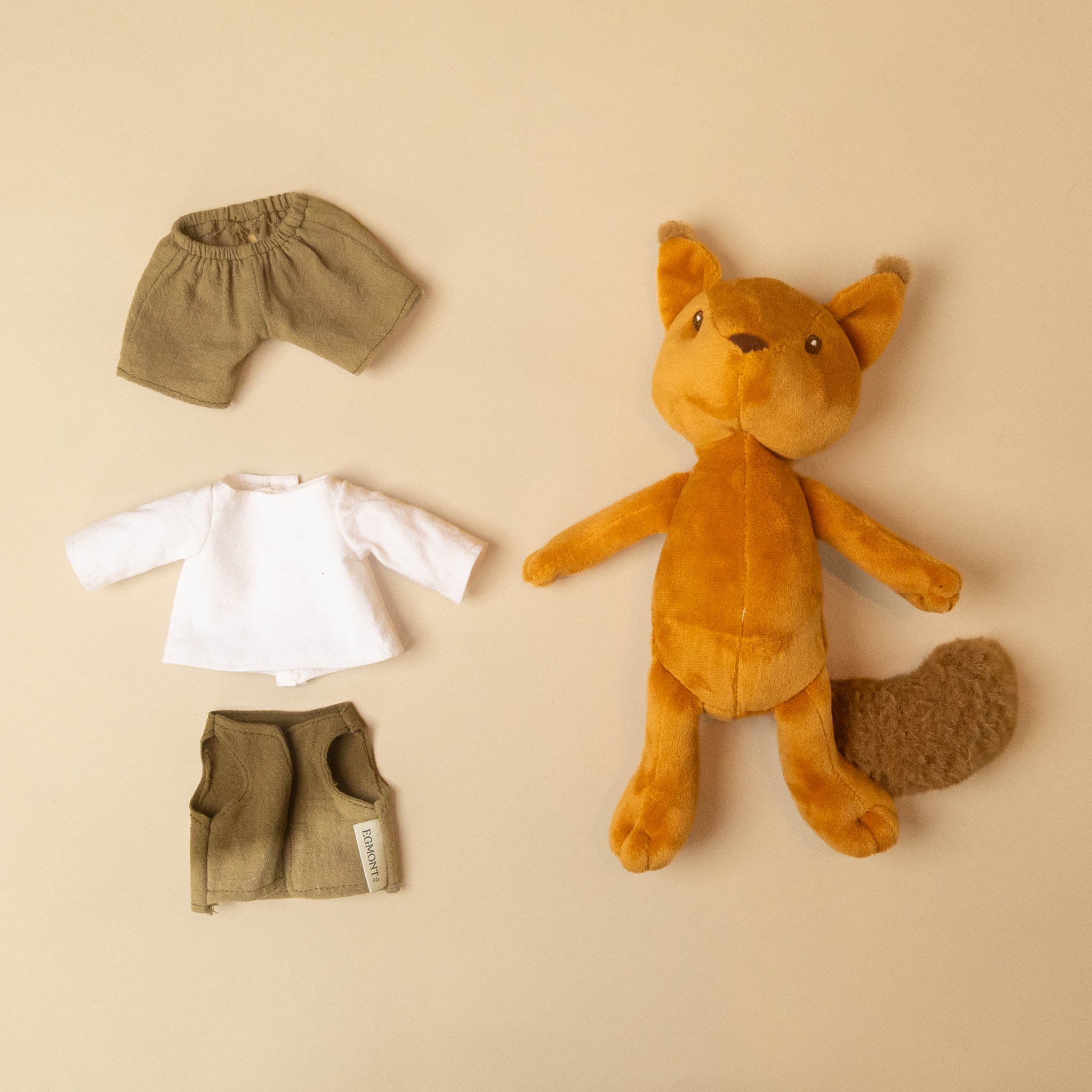 woodland-friends-squirrel-shirt-vest-and-plants with a brown tail is arranged on a warm beige background alongside a matching outfit set: olive green shorts, a white long-sleeve top, and olive green pants.