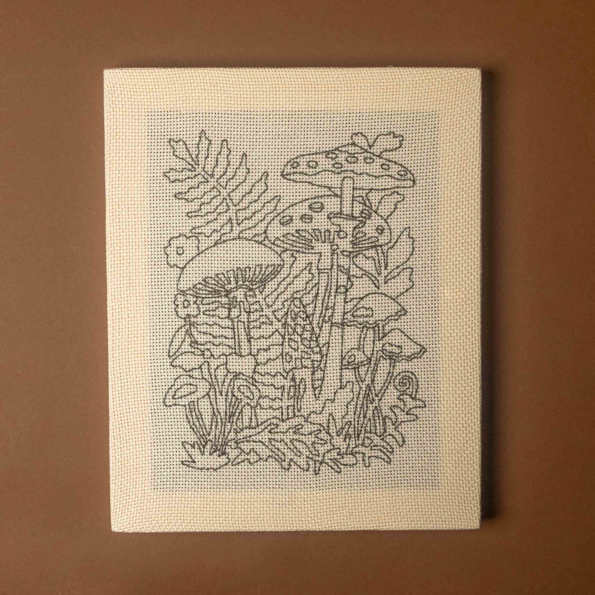 A square canvas art piece featuring a black line drawing of mushrooms and small plants on a light gray-blue grid background, mounted on a cream fabric texture and displayed against a solid brown background.