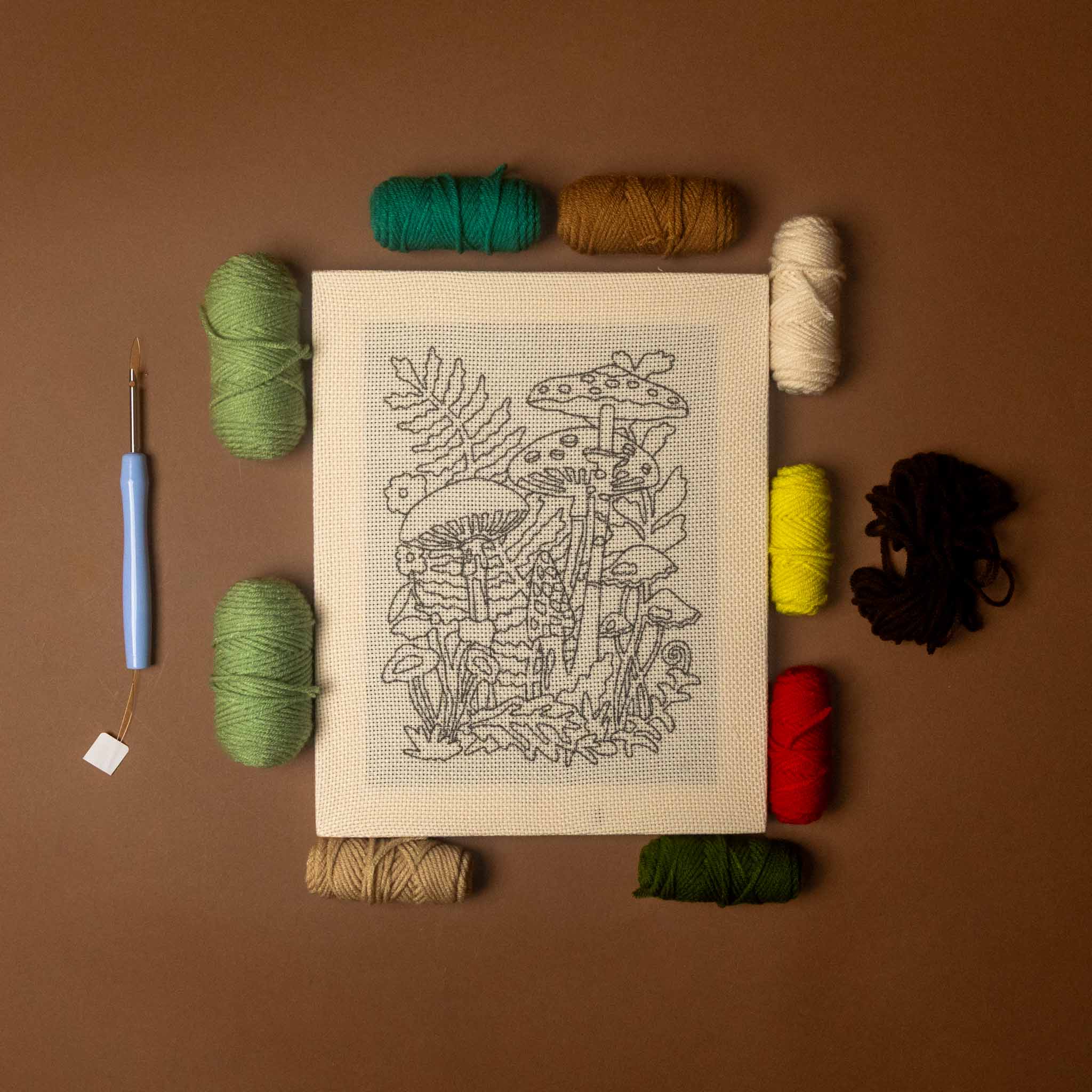 A flat-lay of a mushroom-themed needlepoint canvas surrounded by various colored yarn skeins and a blue-handled crochet hook on a solid brown background.
