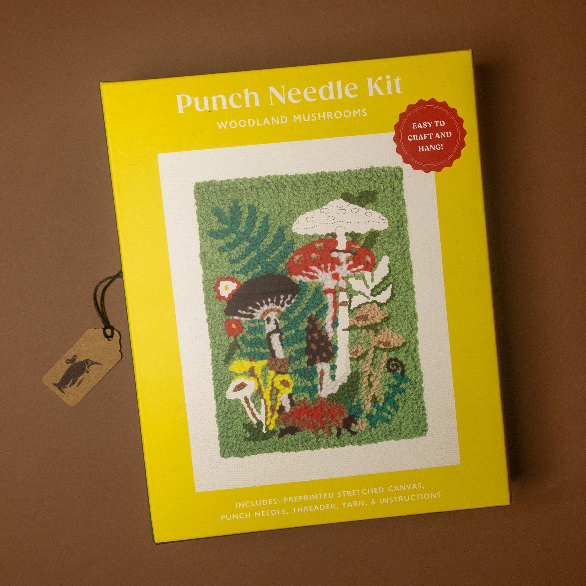 A Punch Needle Kit box for 'Woodland Mushrooms' shown on a brown background with a small hanging tag. The box is bright yellow with a central sample patch showing colorful mushrooms and green foliage, and a red circular badge that says 'Easy to craft and mend!'.