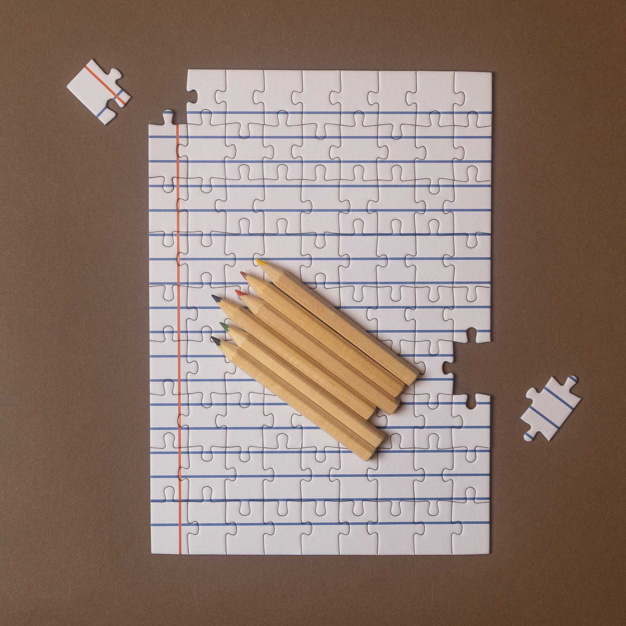 A square jigsaw puzzle printed with notebook paper lines and a red margin line, arranged on a brown background with five wooden pencils on top and two loose puzzle pieces nearby.