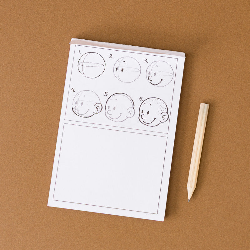 A white sketch pad with black line drawings of cartoon faces, shown on a brown background next to a wooden drawing stick.