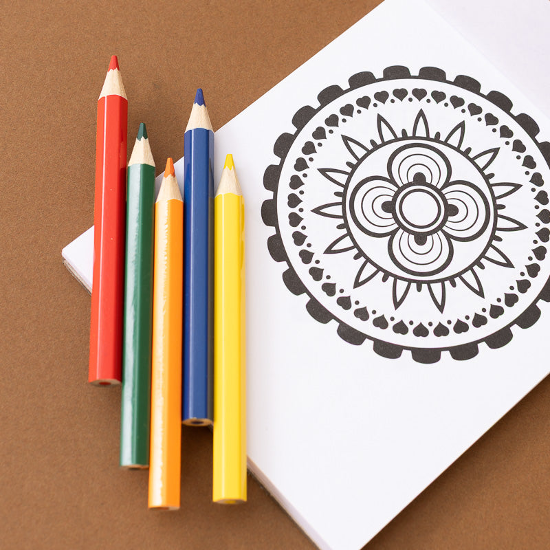 A white coloring book or sheet with a black mandala-style floral line drawing, arranged on a brown surface with five colored pencils in red, green, orange, blue, and yellow leaning against it.