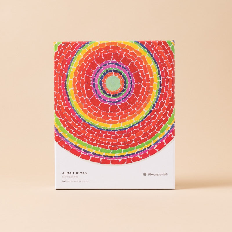 A completed circular mosaic puzzle in a white box, featuring concentric rings of red, orange, yellow, green, purple, and blue brick-like shapes. The box is labeled 'ALMA THOMAS' 500 piece set against a solid beige background.