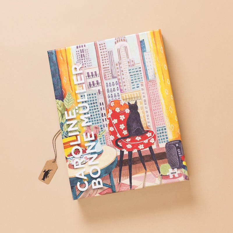 A hardcover notebook titled 'Caroline Bonemuster' featuring a colorful illustration of a black cat sitting on a red floral armchair in front of a city skyline. The notebook is displayed at an angle on a solid beige background with a small paper tag attached.