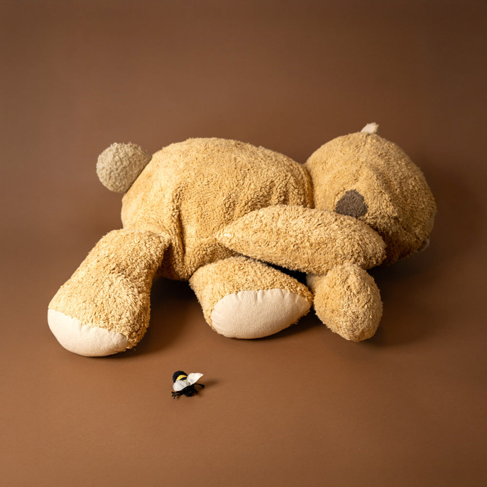 Sleepy-Bear-Floor-Cushion Brown teddy bear lying on a brown surface with a small black and white bee nearby.