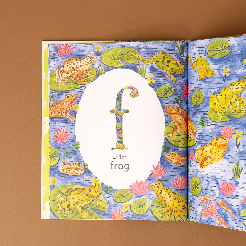 An open children's book showing a frog-themed page with the letter 'f' and the word 'for frog' centered on a blue water background with illustrated frogs, lily pads, and pink flowers.