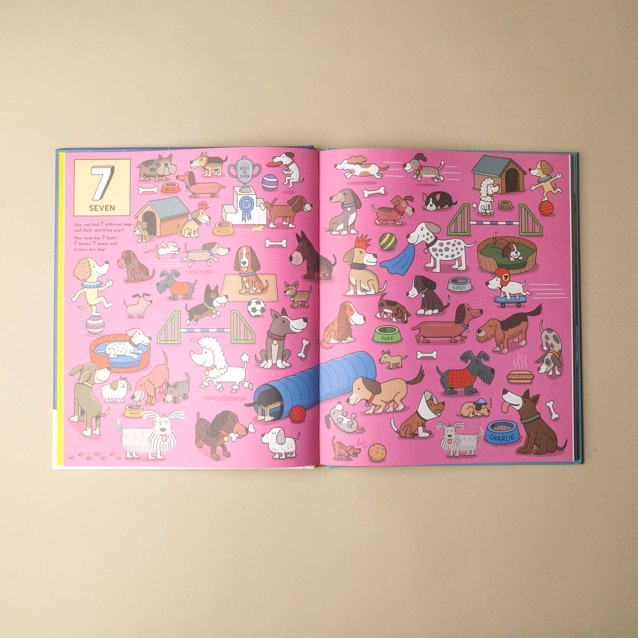An open children's activity book showing a pink page with a cartoon dog-themed puzzle and a yellow '7 SEVEN' box on the left page, photographed on a beige background.