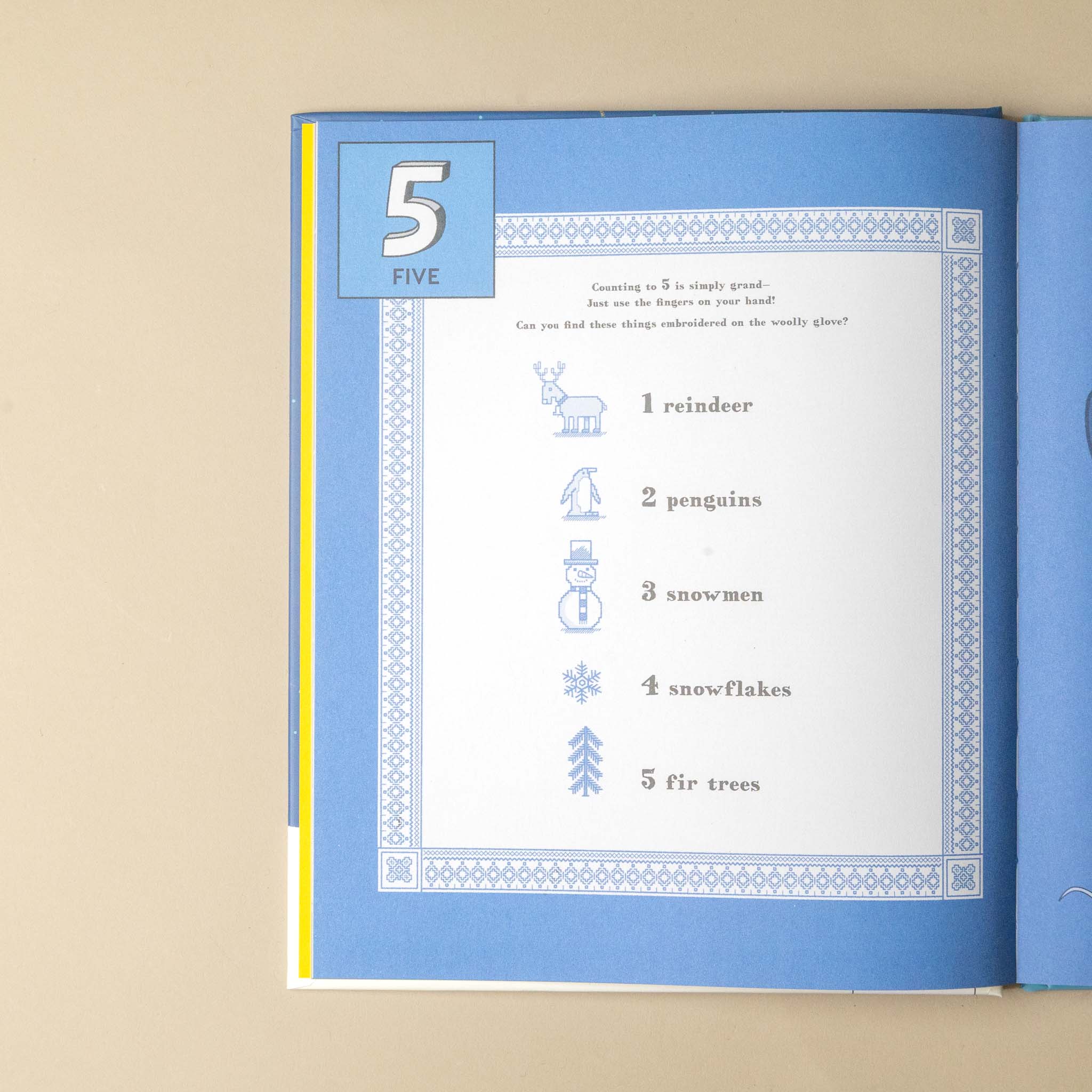 An open children's book or activity booklet on a beige surface, showing a blue cover and an interior page with a counting activity titled '5 FIVE' and simple winter illustrations and text.