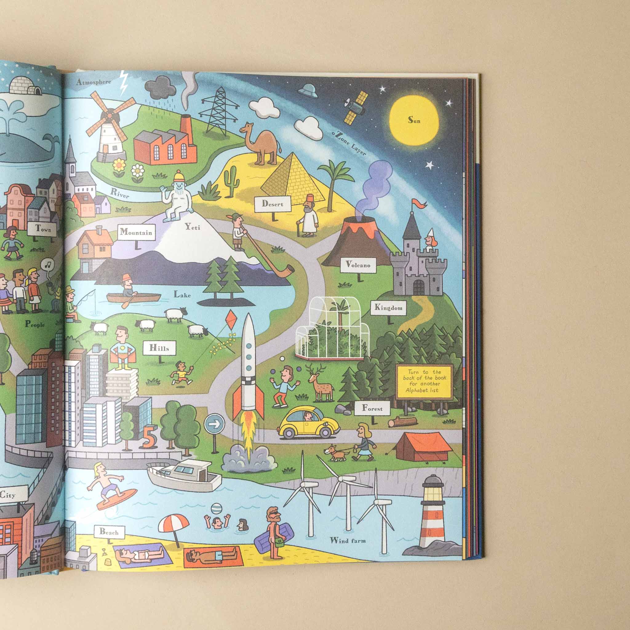 An open children's illustrated book spread on a beige surface, showing a colorful cartoon map with labeled places, a rocket, a lighthouse, a castle, a windmill, and other whimsical scenes.