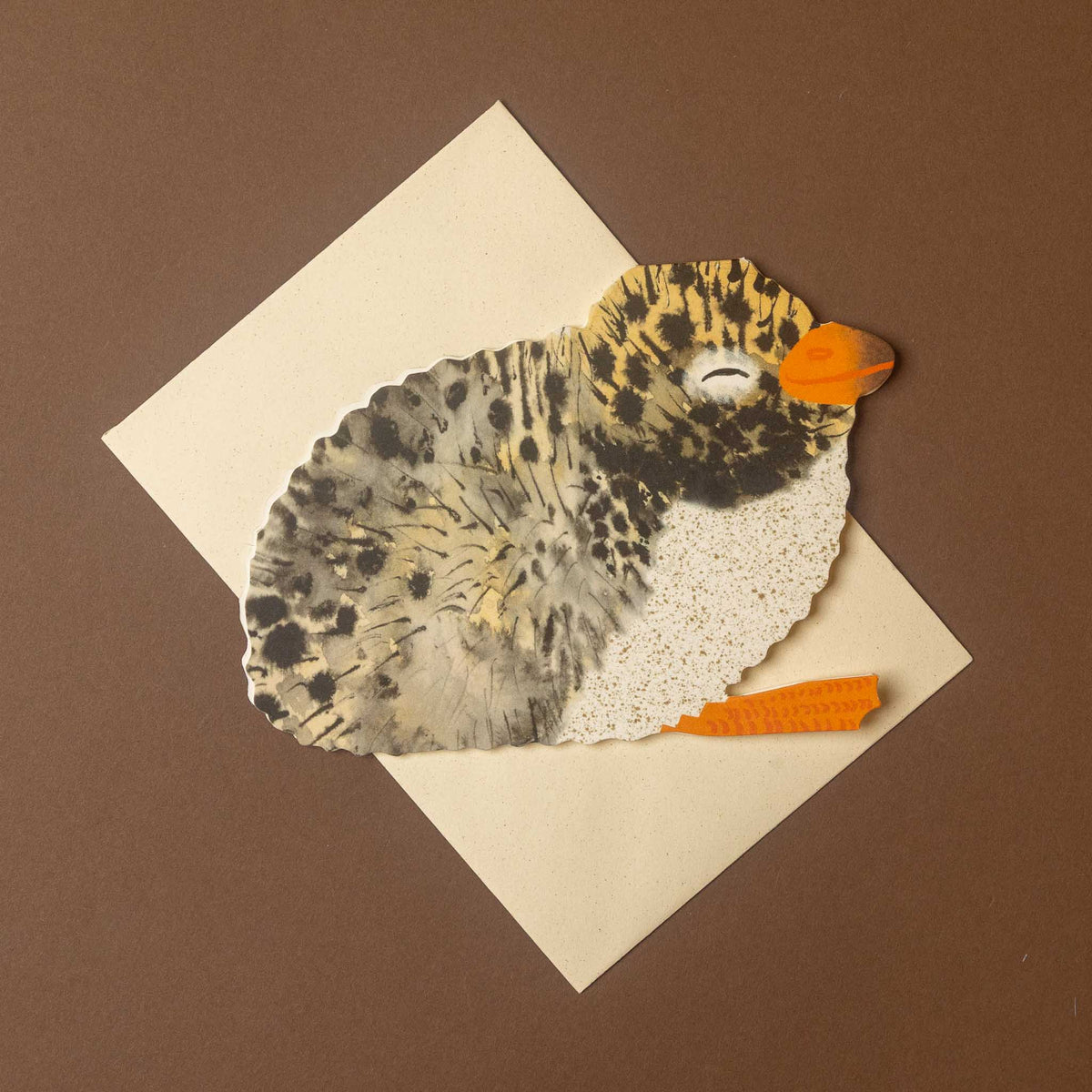 arctic-tern-chick-greeting-card on a cream paper envelope against a solid brown background. The bird features textured gray and black feathers, a speckled cream body, and bright orange beak and tail piece.