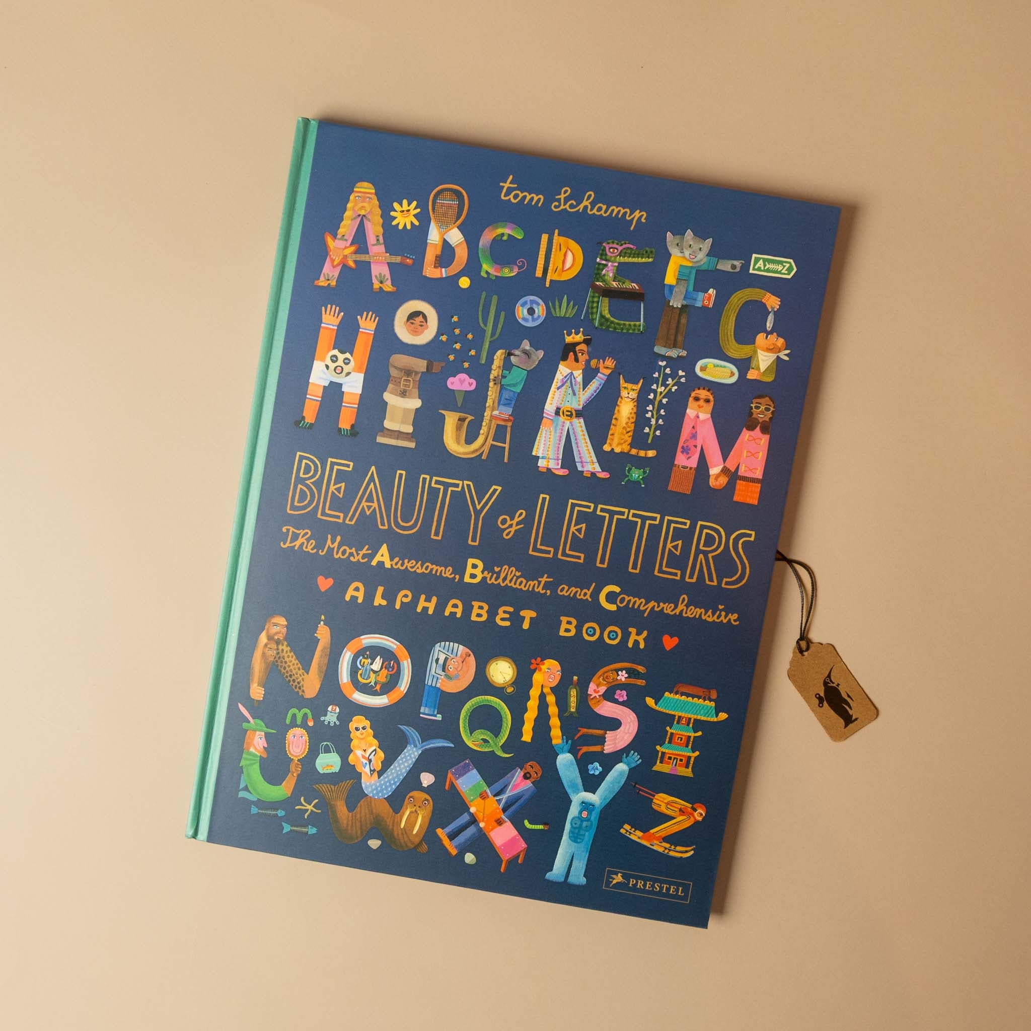 A children's alphabet book titled 'Beauty of Letters' by Tom Schamp, featuring a dark blue cover decorated with colorful illustrated letters and characters, photographed on a warm beige background with a small wooden tag attached.