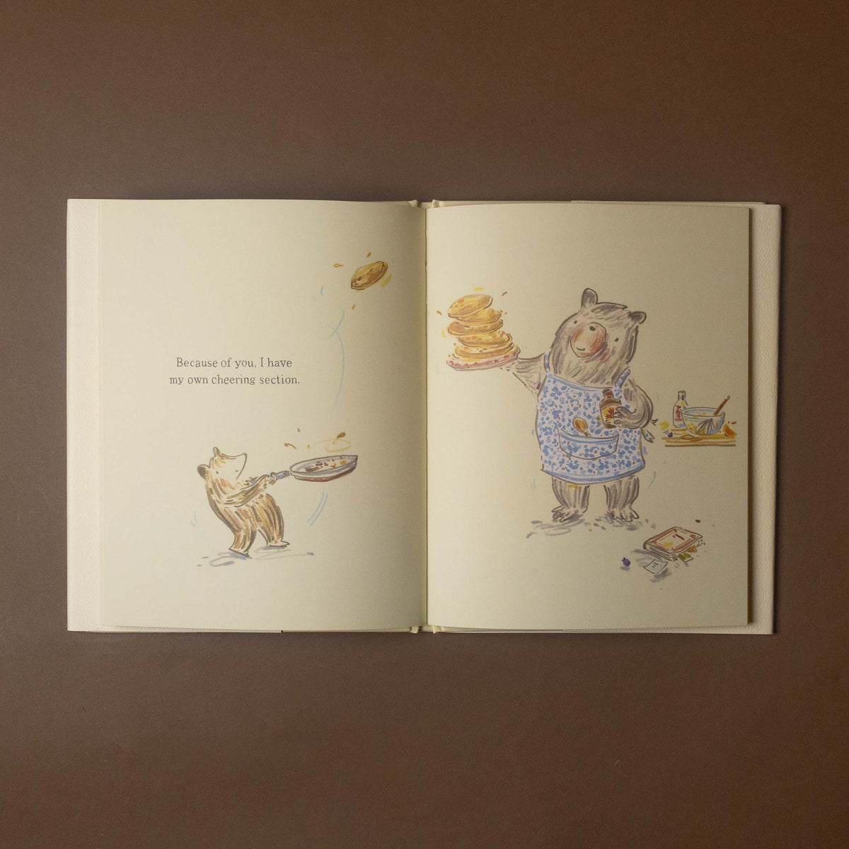 An open illustrated book laid flat on a brown background. The pages show whimsical animal drawings: a cat throwing a pan and a bear wearing a blue patterned apron holding pancakes.
