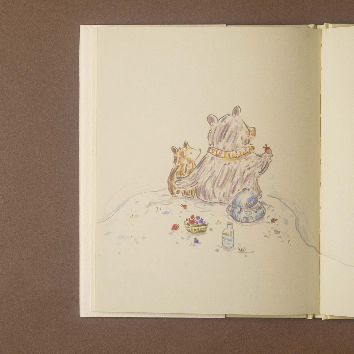 An open sketchbook or art journal on a brown background, featuring a watercolor illustration of two bears sitting on a hill with small scattered details like berries, a bottle, and a blue hat.