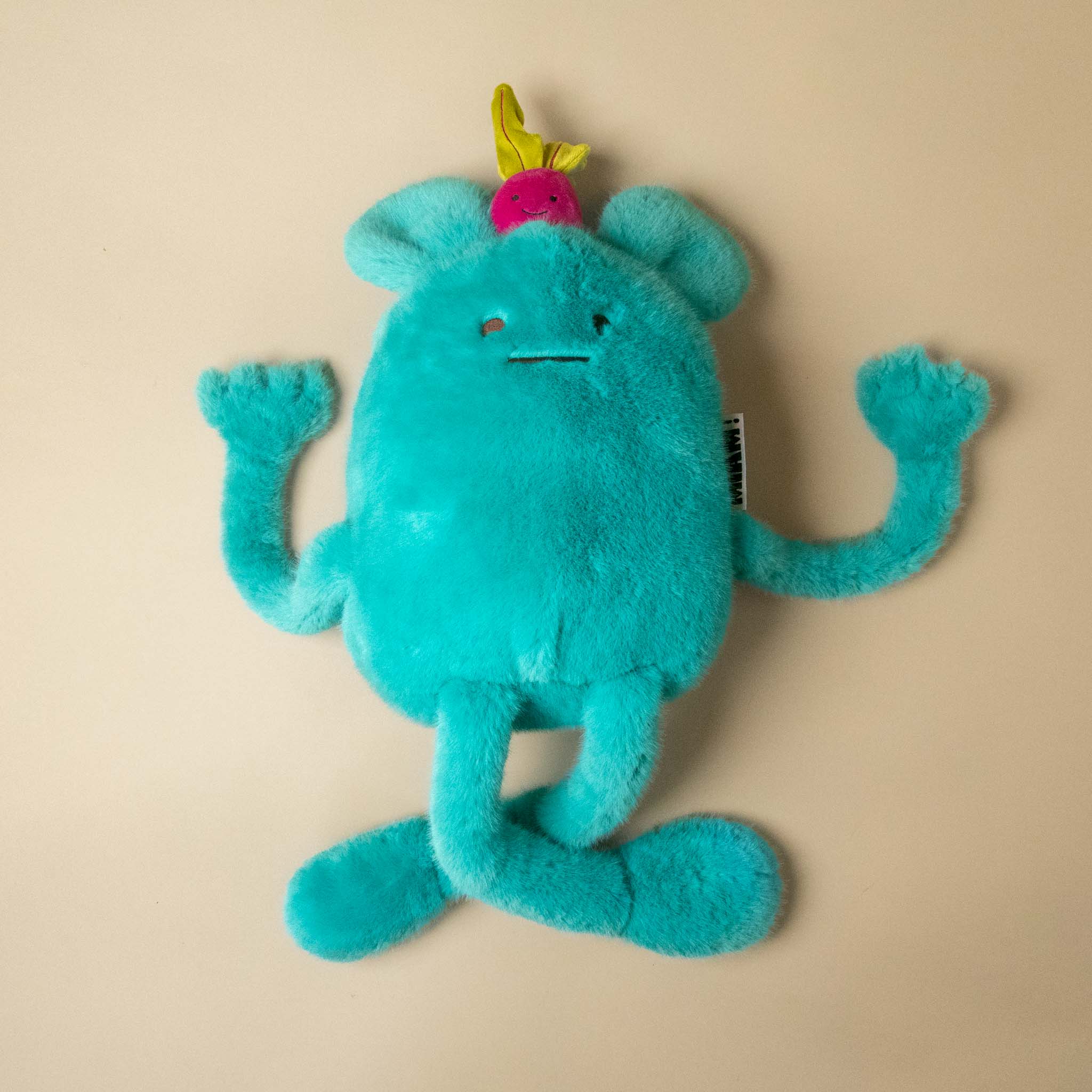 A turquoise blue plush billie-radish-grand toy with long limbs, a small pink flower on its head, and a hangtag, photographed on a plain beige background.