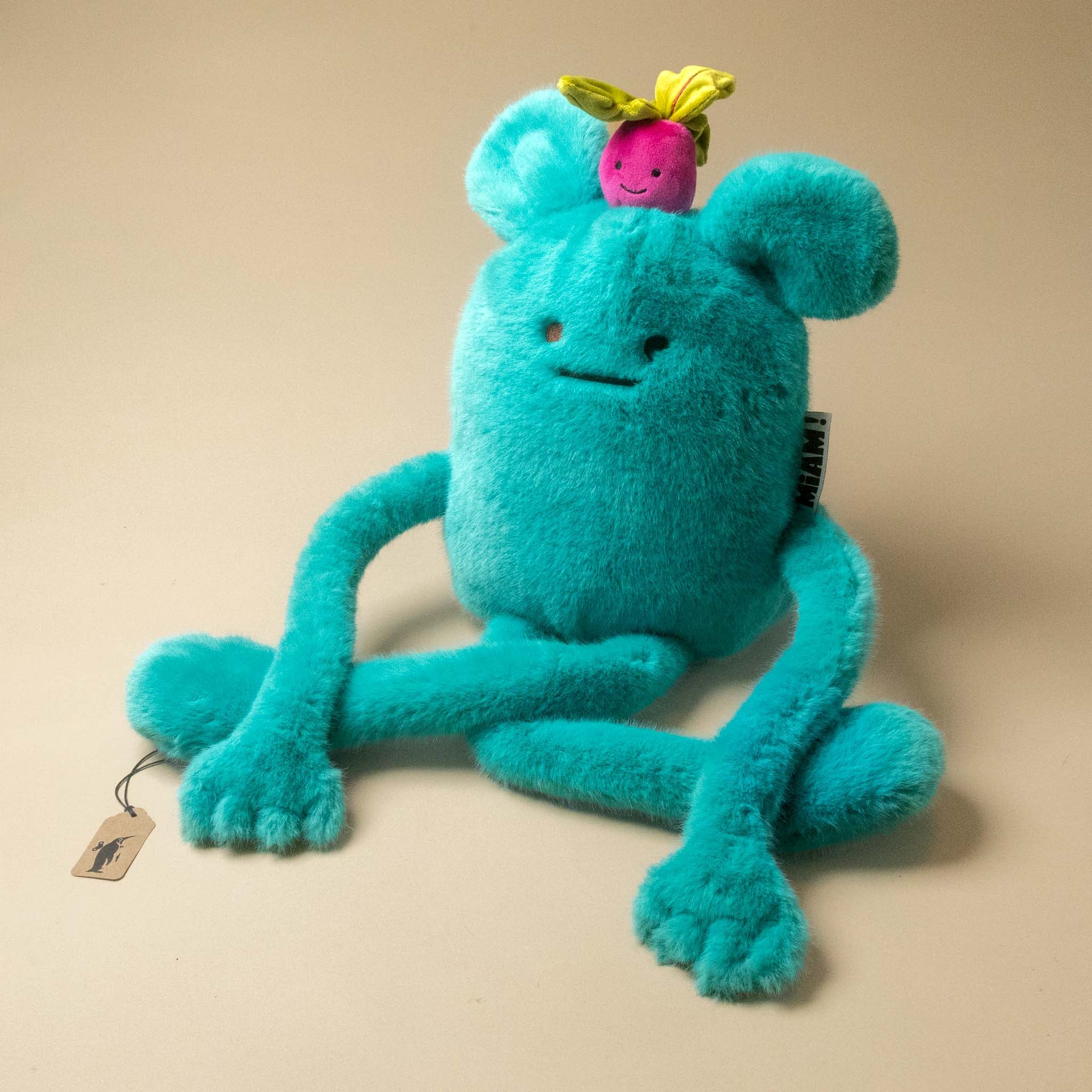 A turquoise blue plush billie-radish-grand toy with long limbs, a small pink flower on its head, and a hangtag, photographed on a plain beige background.