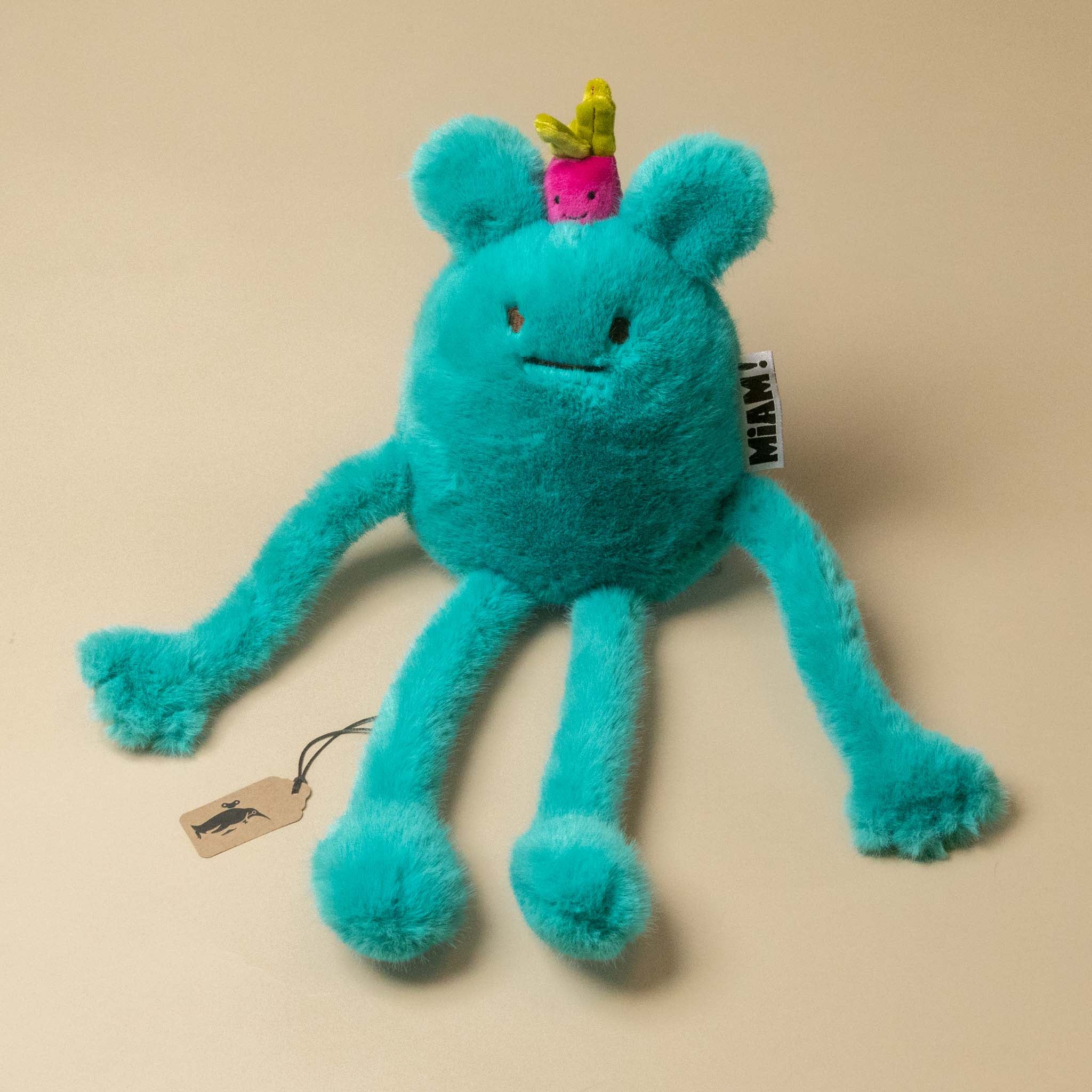 A turquoise blue fuzzy A turquoise blue plush billie-radish-petit toy with long limbs, a small pink flower on its head, and a hangtag, photographed on a plain beige background.plush toy with a small pink and green radish  on its head, sitting on a plain beige background. 