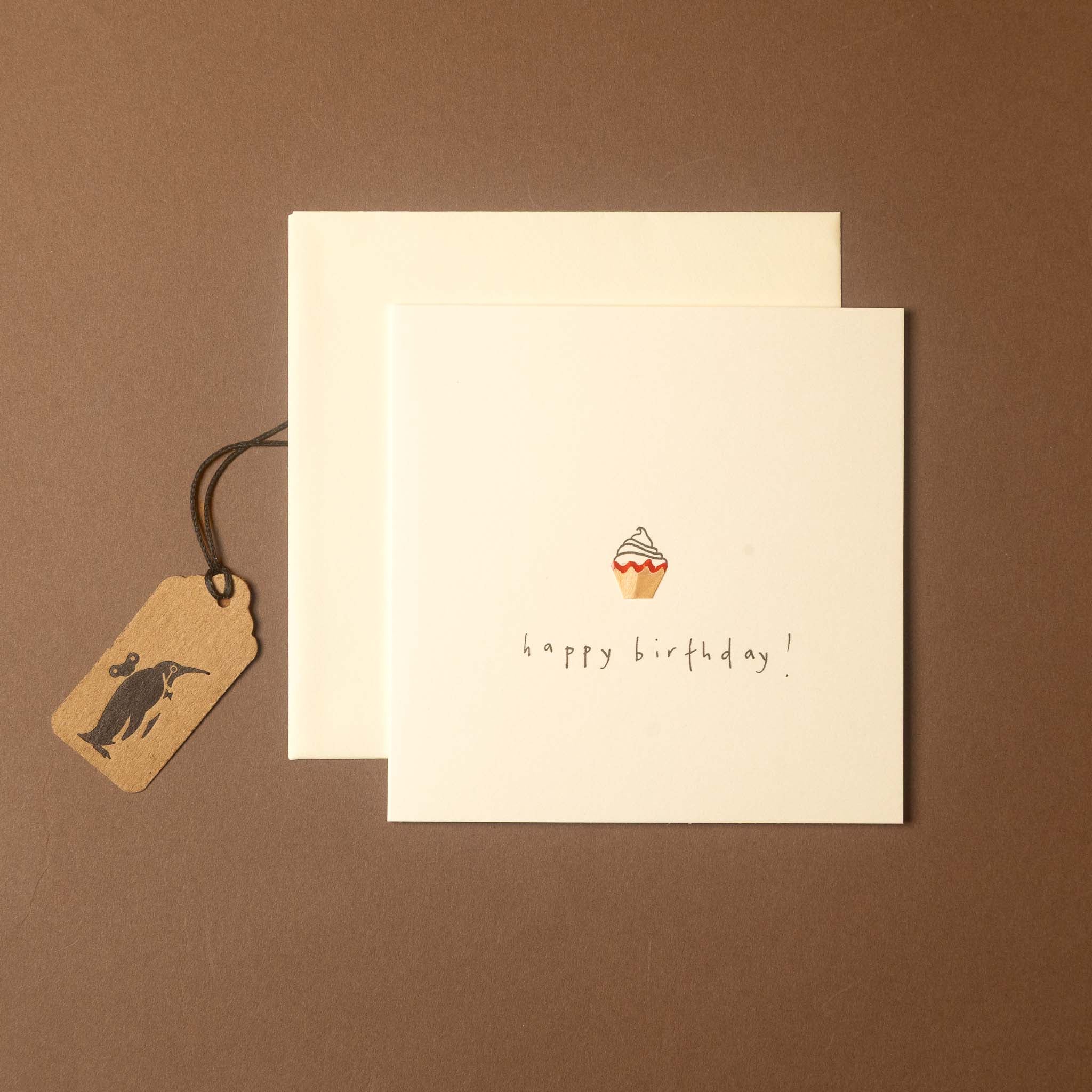 A square birthday card with a small cupcake illustration and the words 'happy birthday!' in black script, shown with a matching cream envelope and a small kraft paper tag on a brown background.