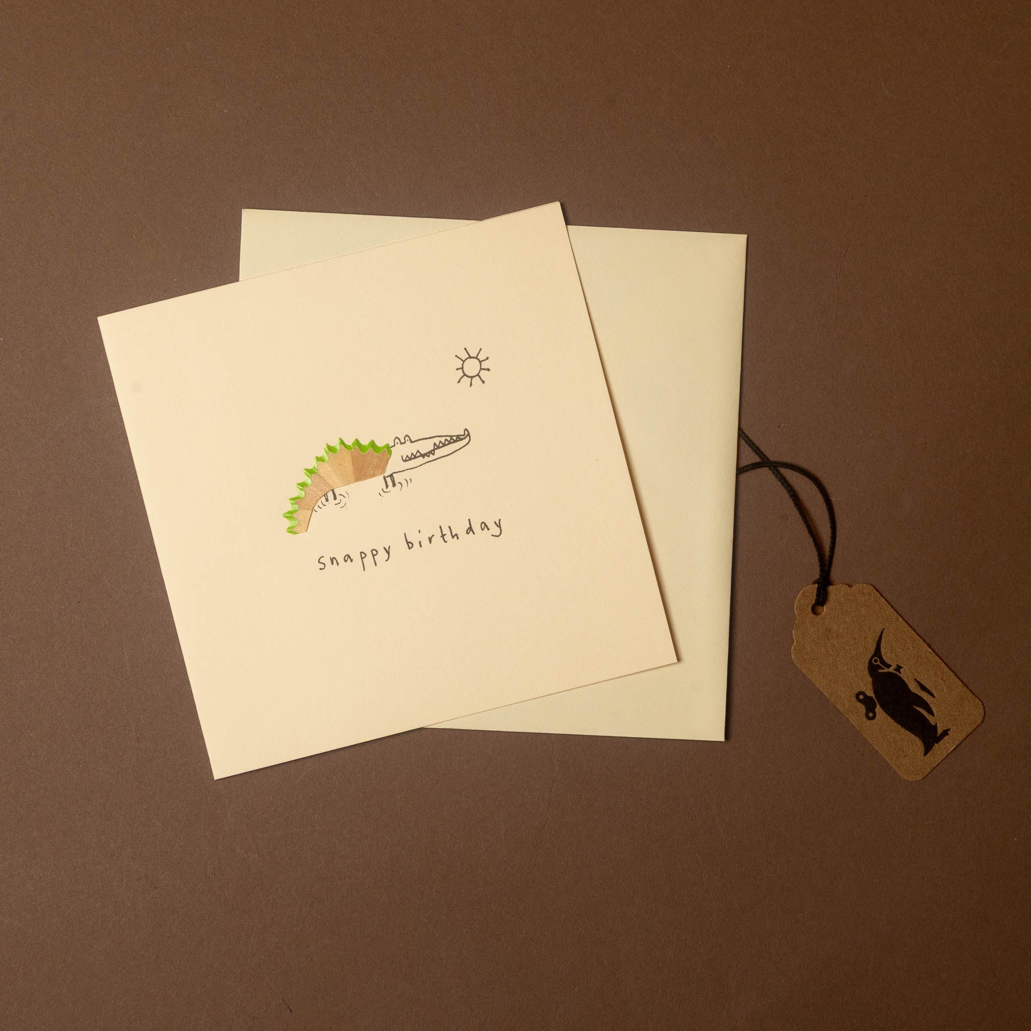 A square cream greeting card with a simple illustration of a smiling shark and the words 'snappy birthday' on a brown background, with a matching cream envelope and a small kraft gift tag tied to it.