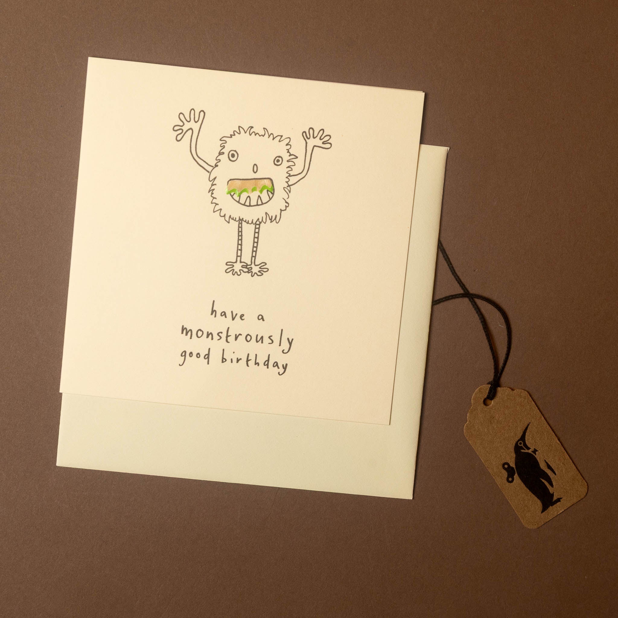 A square birthday card with a hand-drawn monster illustration and the text 'have a monstrously good birthday,' shown with an envelope and a brown gift tag on a brown background.